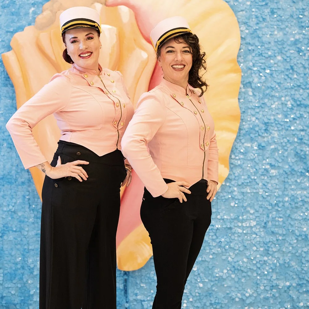 Two women dressed in vintage flight attendant uniforms with pink tops, black pants, and white hats with gold accents, standing in front of a colorful backdrop featuring a large seashell and a blue textured background.
