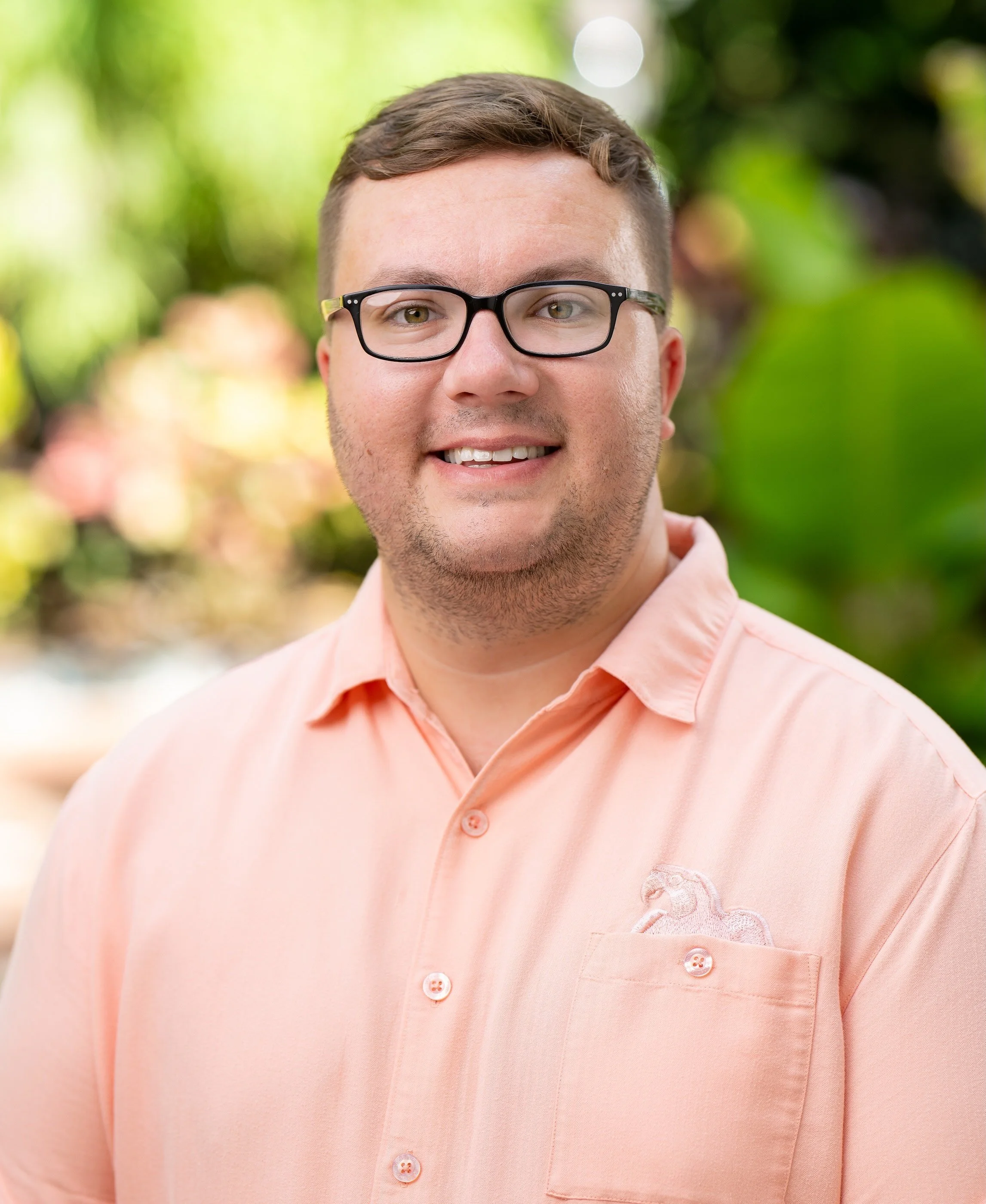 A young man with short brown hair, glasses, and a light beard, wearing a peach-colored shirt, standing outdoors with blurred green foliage in the background.