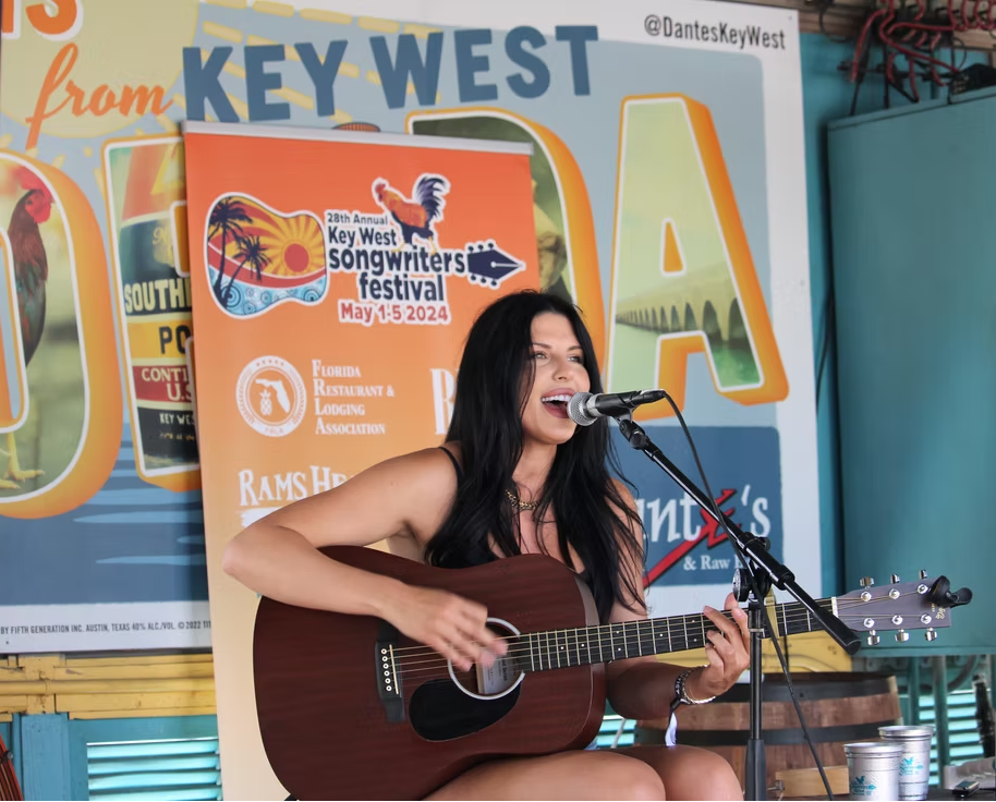 A woman with long dark hair performs with an acoustic guitar on stage at the Key West Songwriters Festival, singing into a microphone.