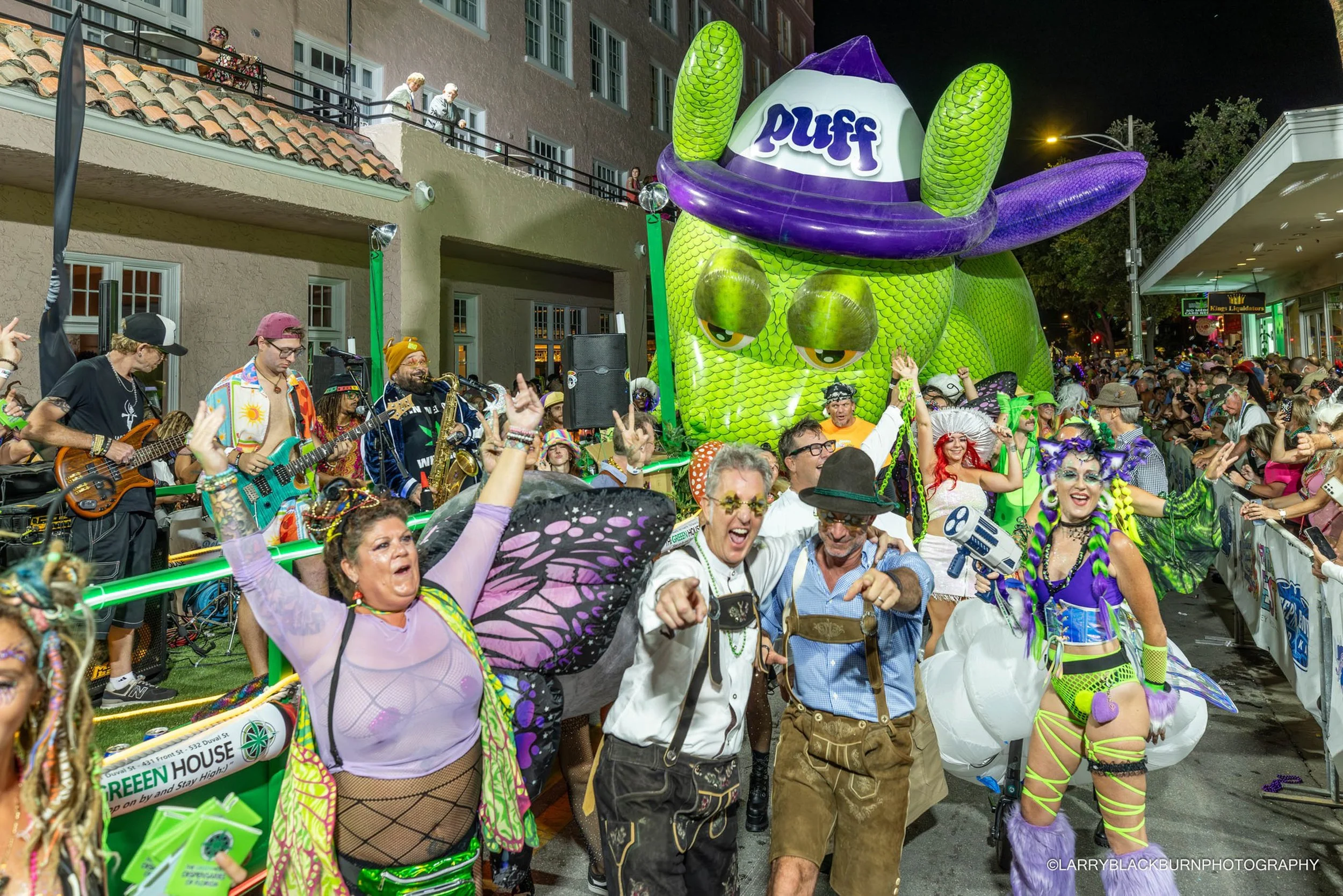 A lively street parade at night with costumed performers, a large inflatable dragon, a band playing, and spectators watching from balconies and sidewalks.