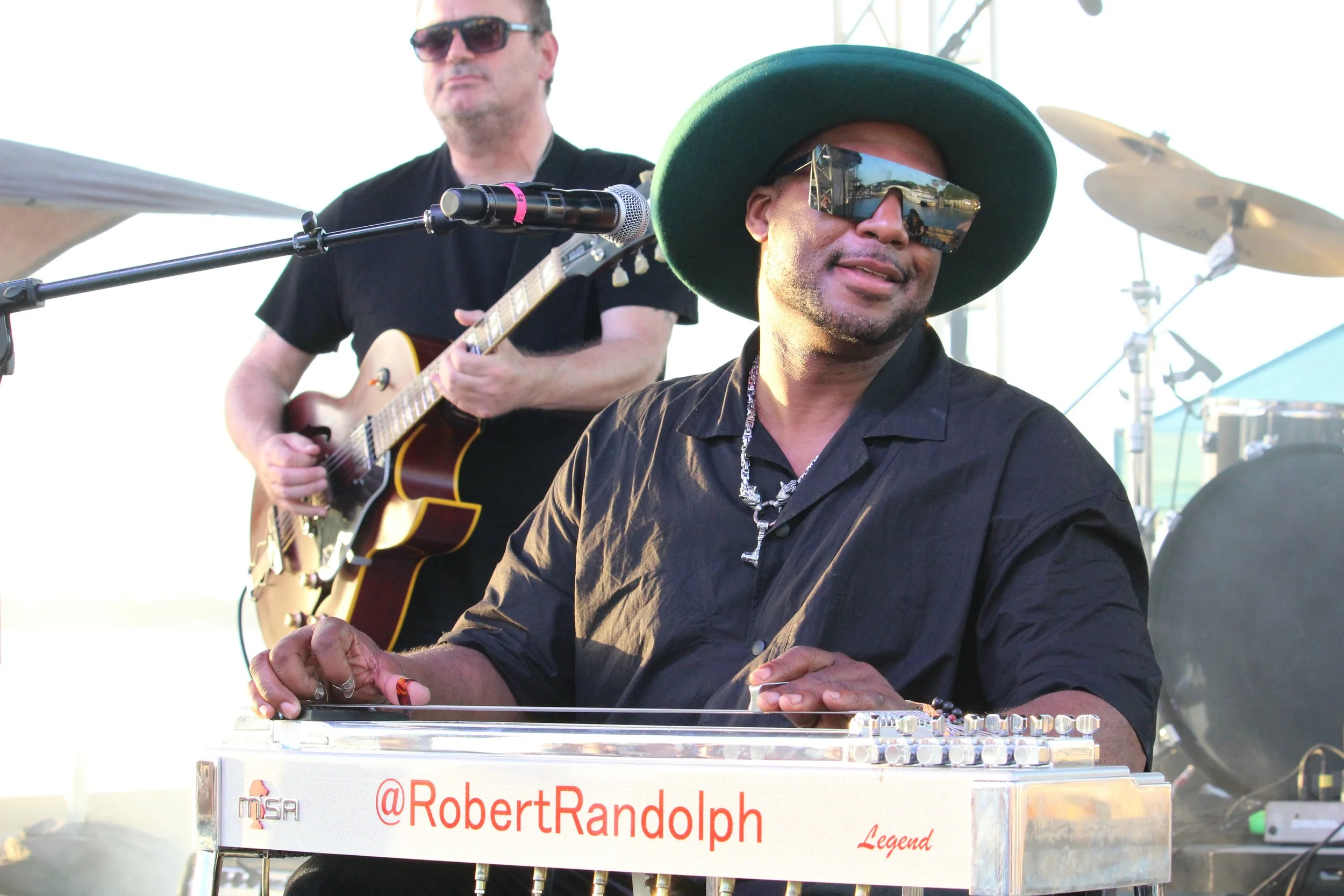 Musician playing a keyboard with a social media handle '@RobertRandolph' visible, wearing a large green hat, sunglasses, and a black shirt, with a guitarist in the background.