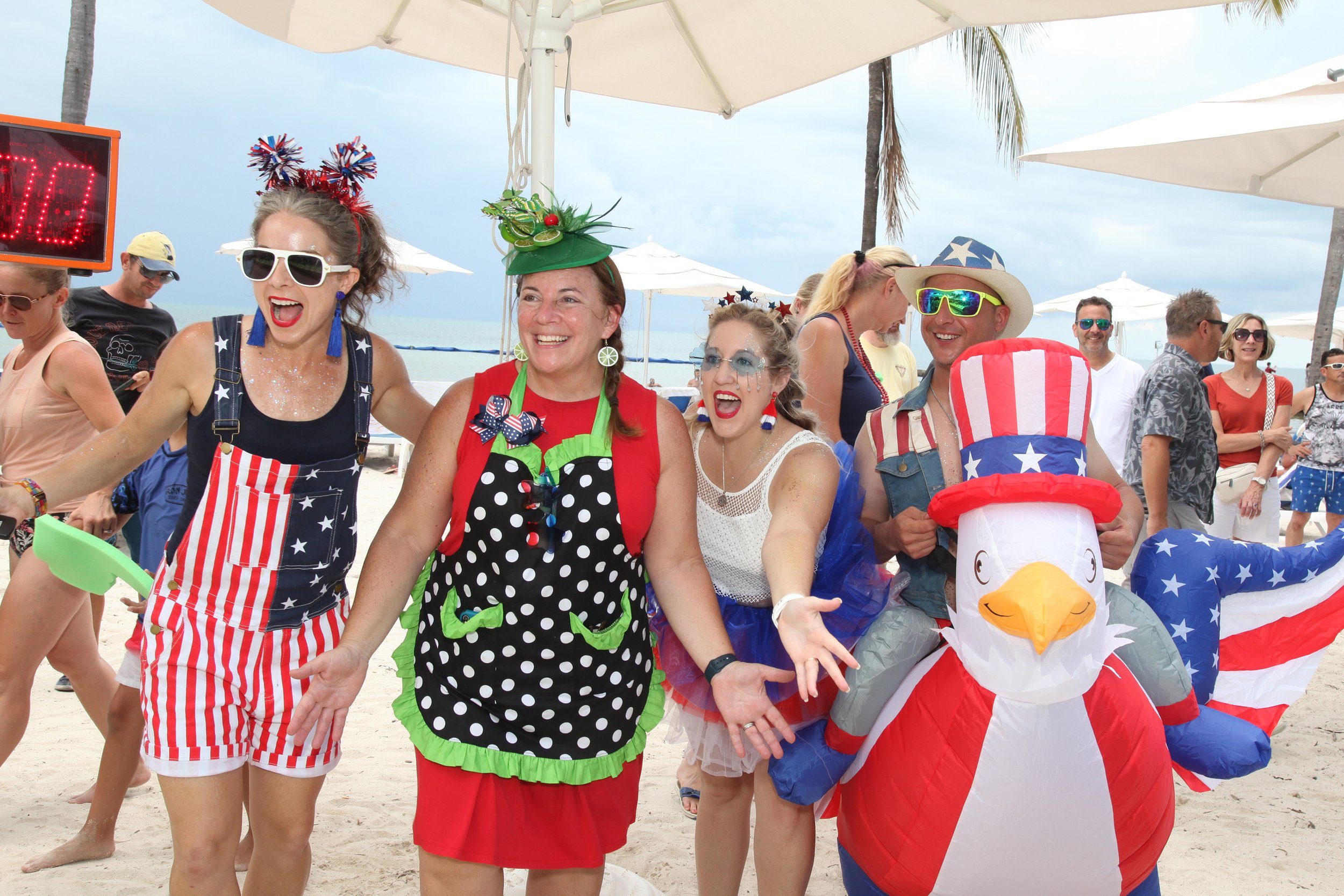 Group of people on the beach dressed in patriotic and festive clothing, celebrating with a large inflatable turkey wearing a Uncle Sam hat, under umbrellas with palm trees in the background.