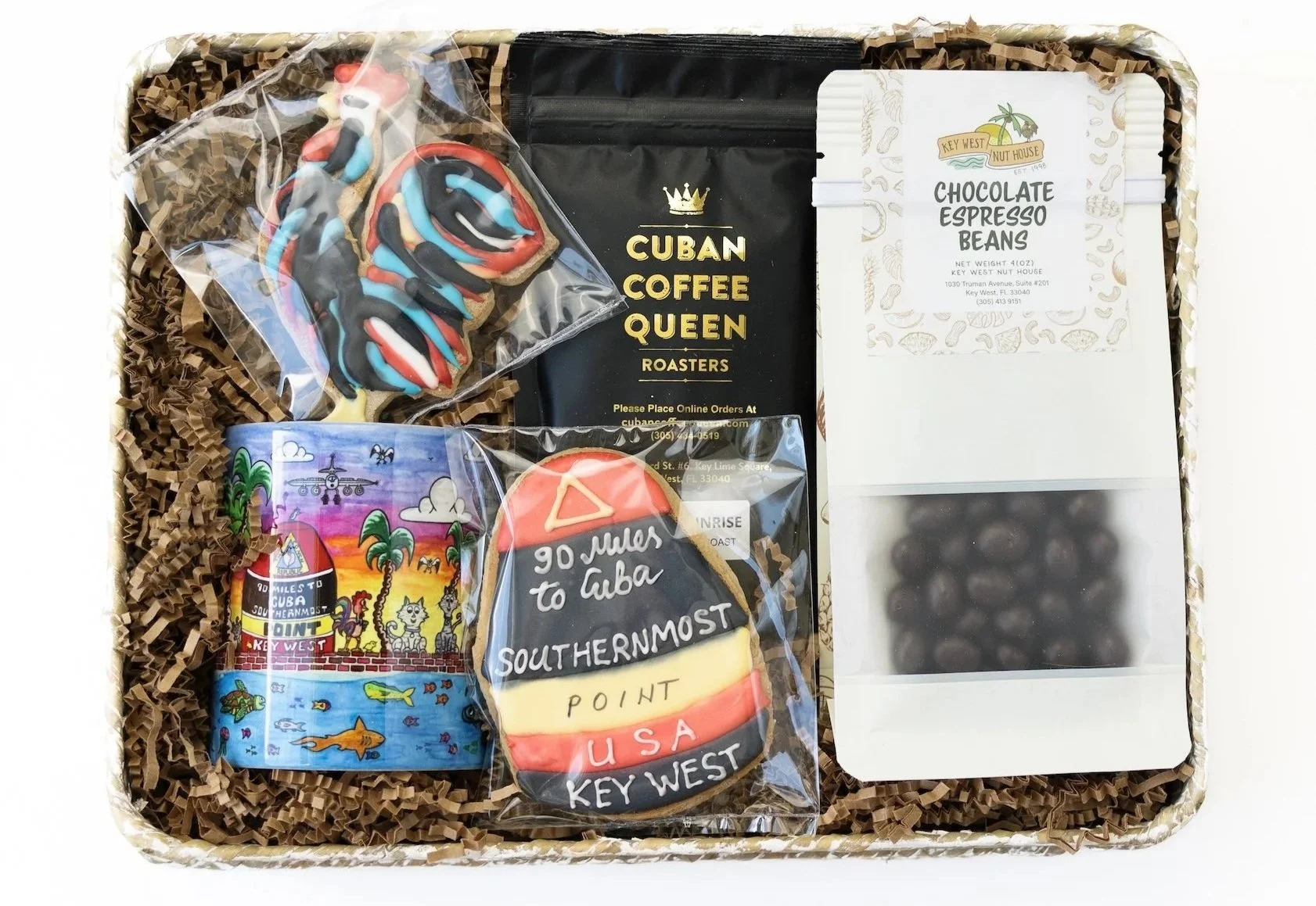 Gift box containing colorful cookies, a packet of Cuban coffee beans, a decorative magnet with Cuban imagery, and a packet of dark chocolate espresso beans, all arranged on crinkled paper filler.