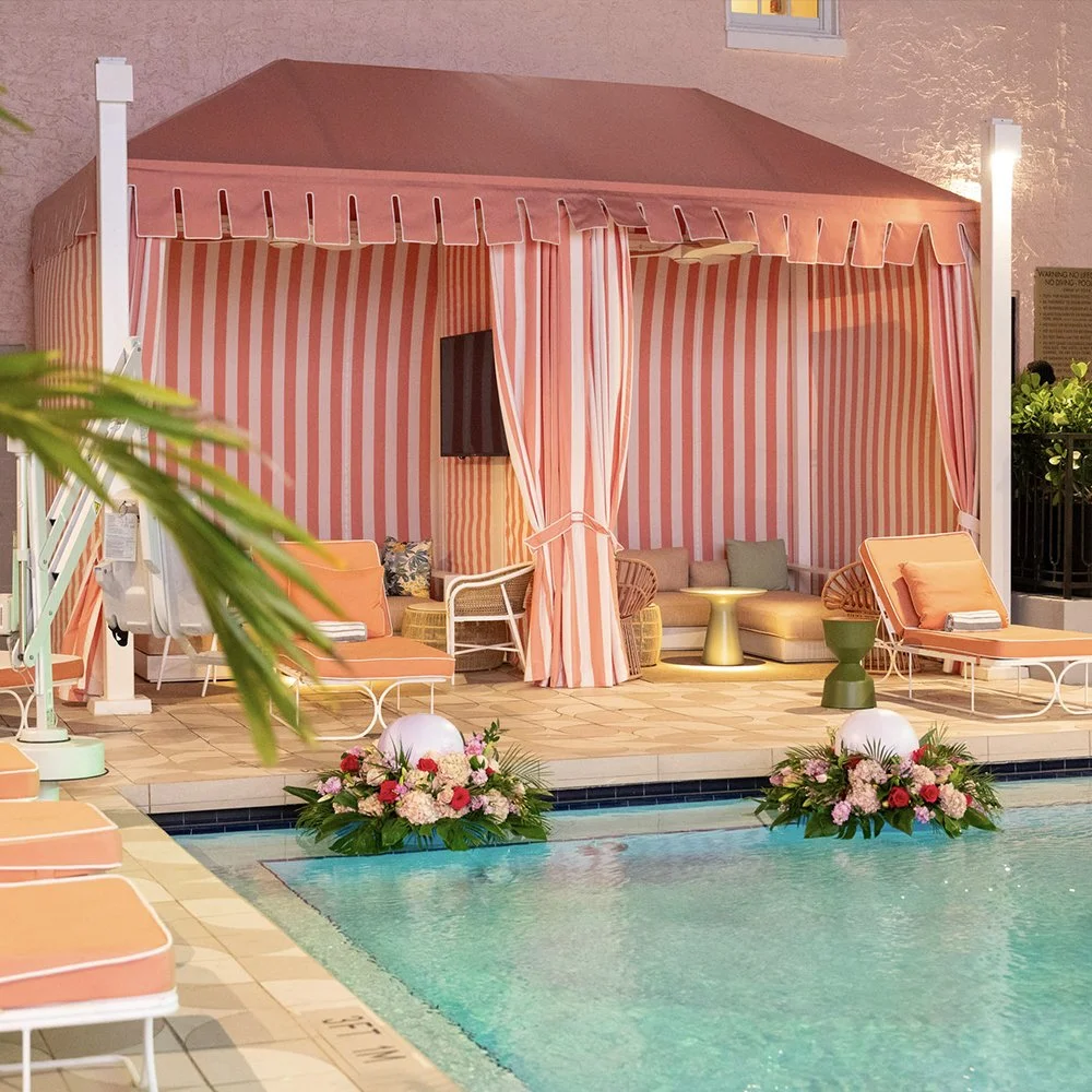 A poolside cabana with pink and white striped curtains, pink cushioned lounge chairs, and floral arrangements at the edge of a swimming pool.