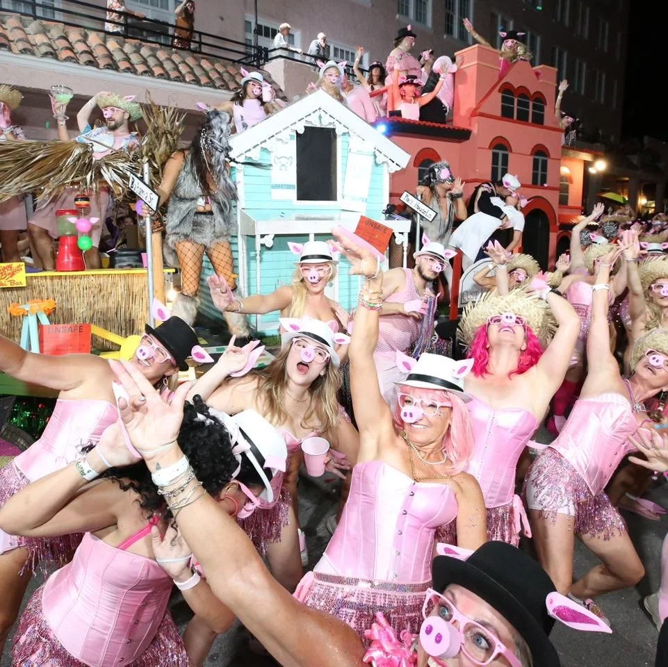 Participants in a lively parade dressed as pigs with pink costumes, pig face masks, and pig ears, dancing and celebrating on the street in front of colorful floats resembling pig-themed houses.