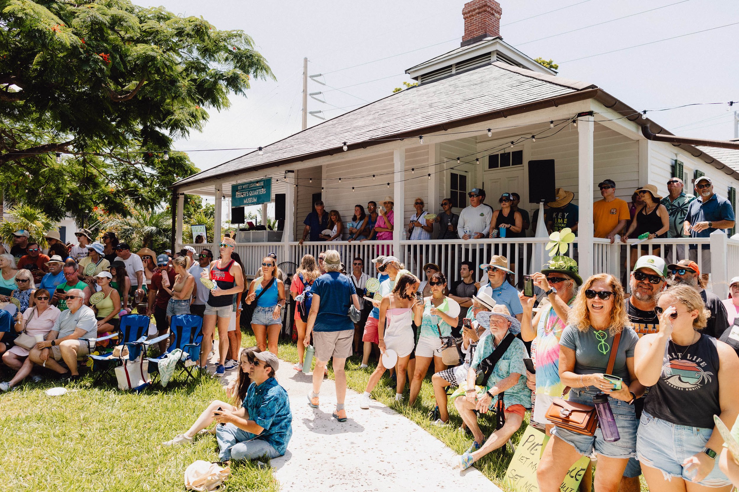 A crowd of people gathered outside a historic white wooden building with a porch and string lights, some seated on chairs and others standing, with trees and greenery in the background.