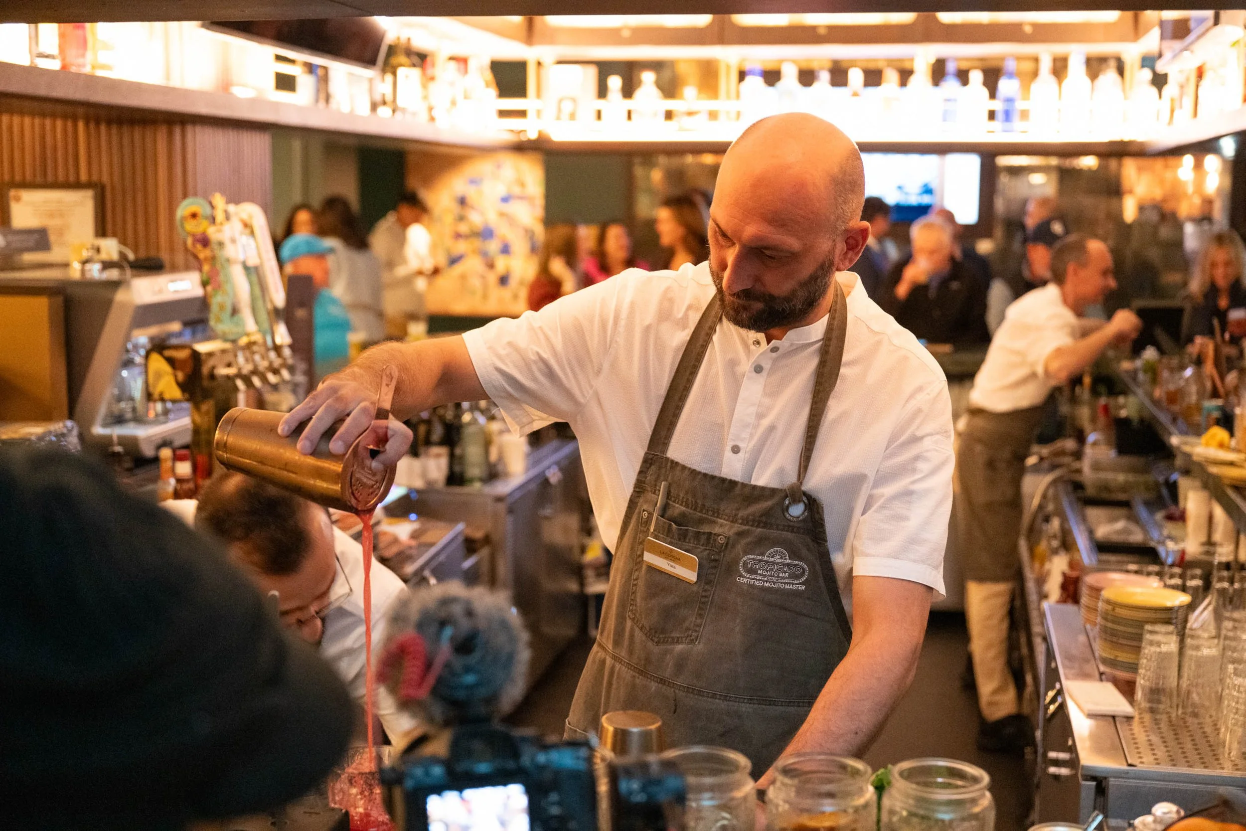 Bald bartender pouring a pink drink into a glass at a busy bar with patrons in the background.