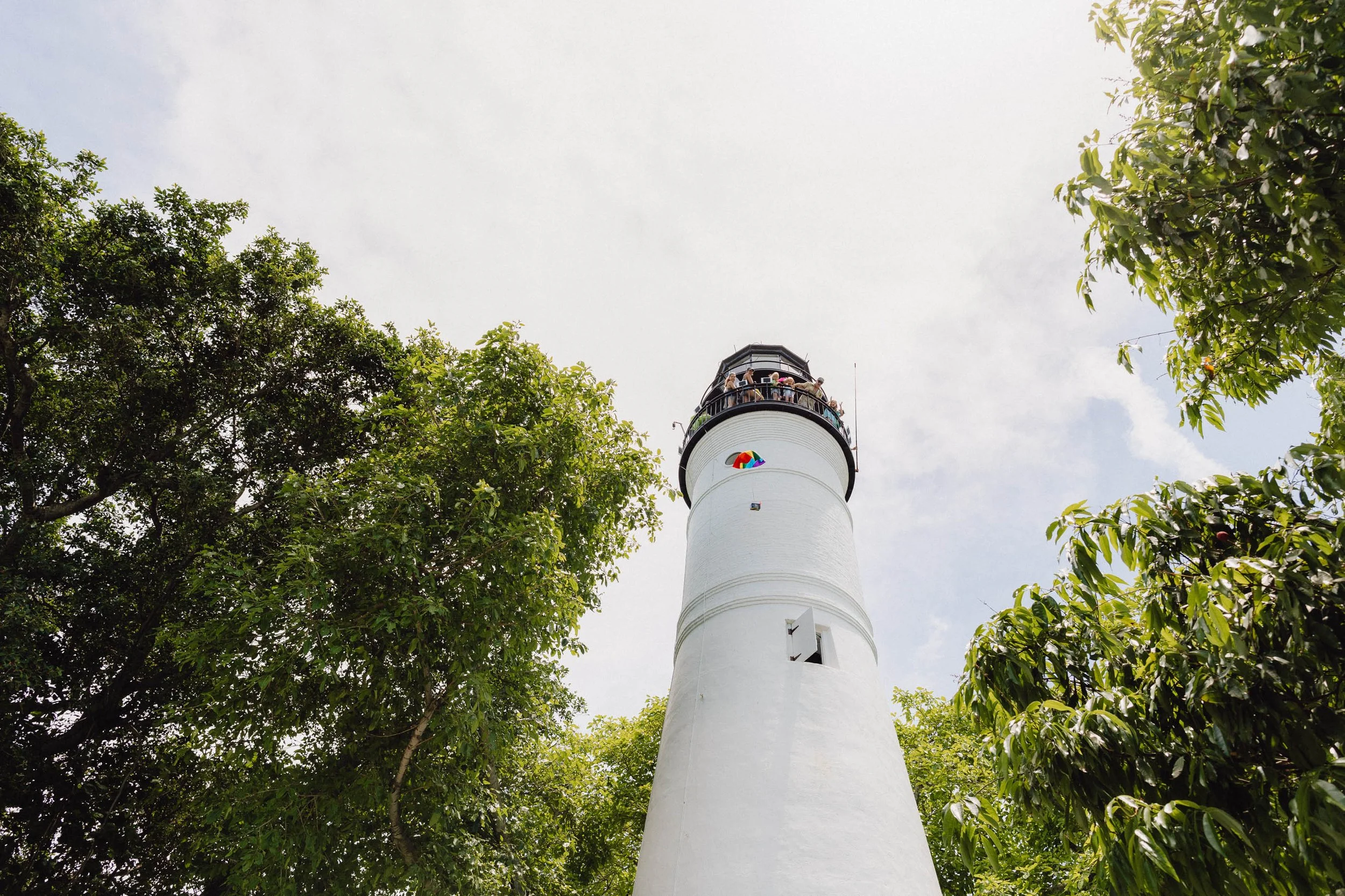 A white lighthouse surrounded by green trees, with people on the top viewing platform under a cloudy sky.