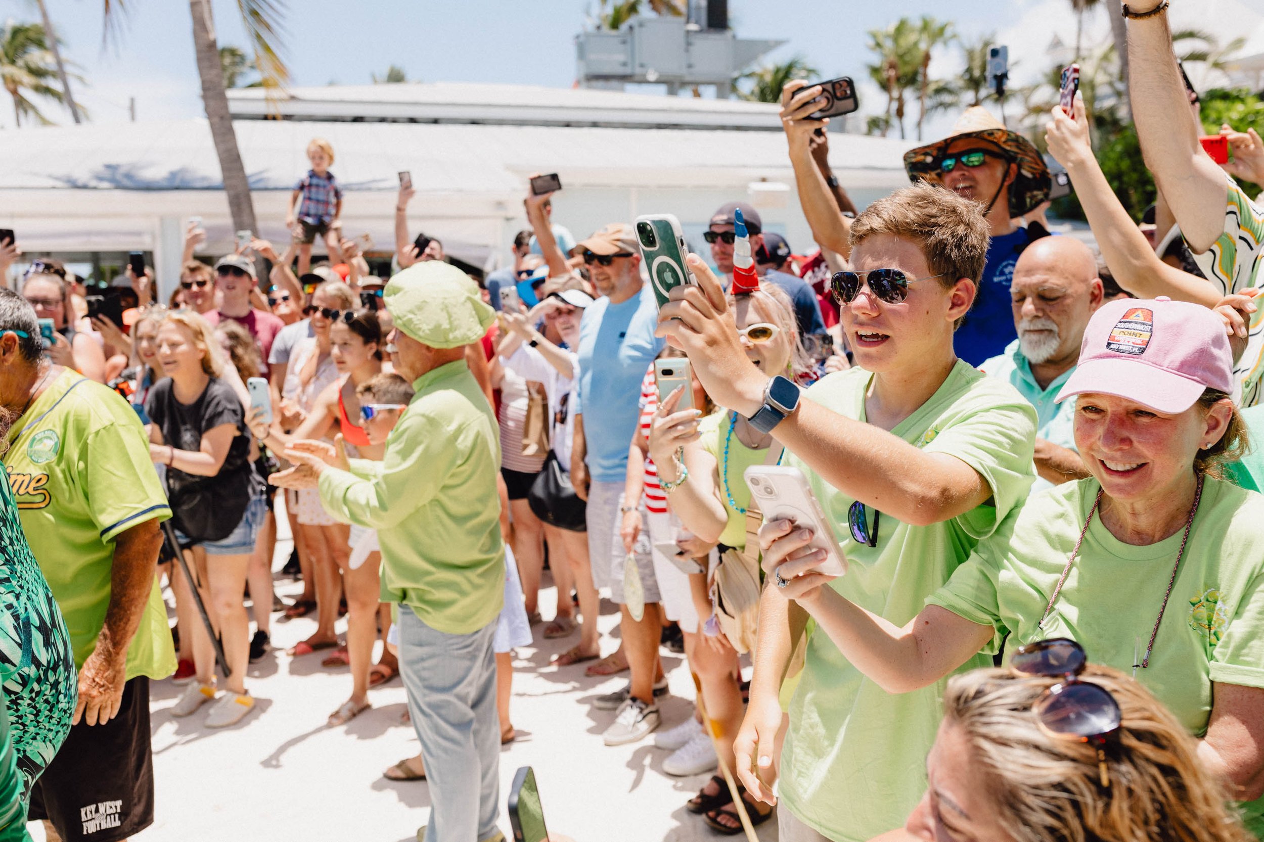 Crowd of people at an outdoor event, many holding phones and taking photos, with palm trees and a white building in the background on a sunny day.