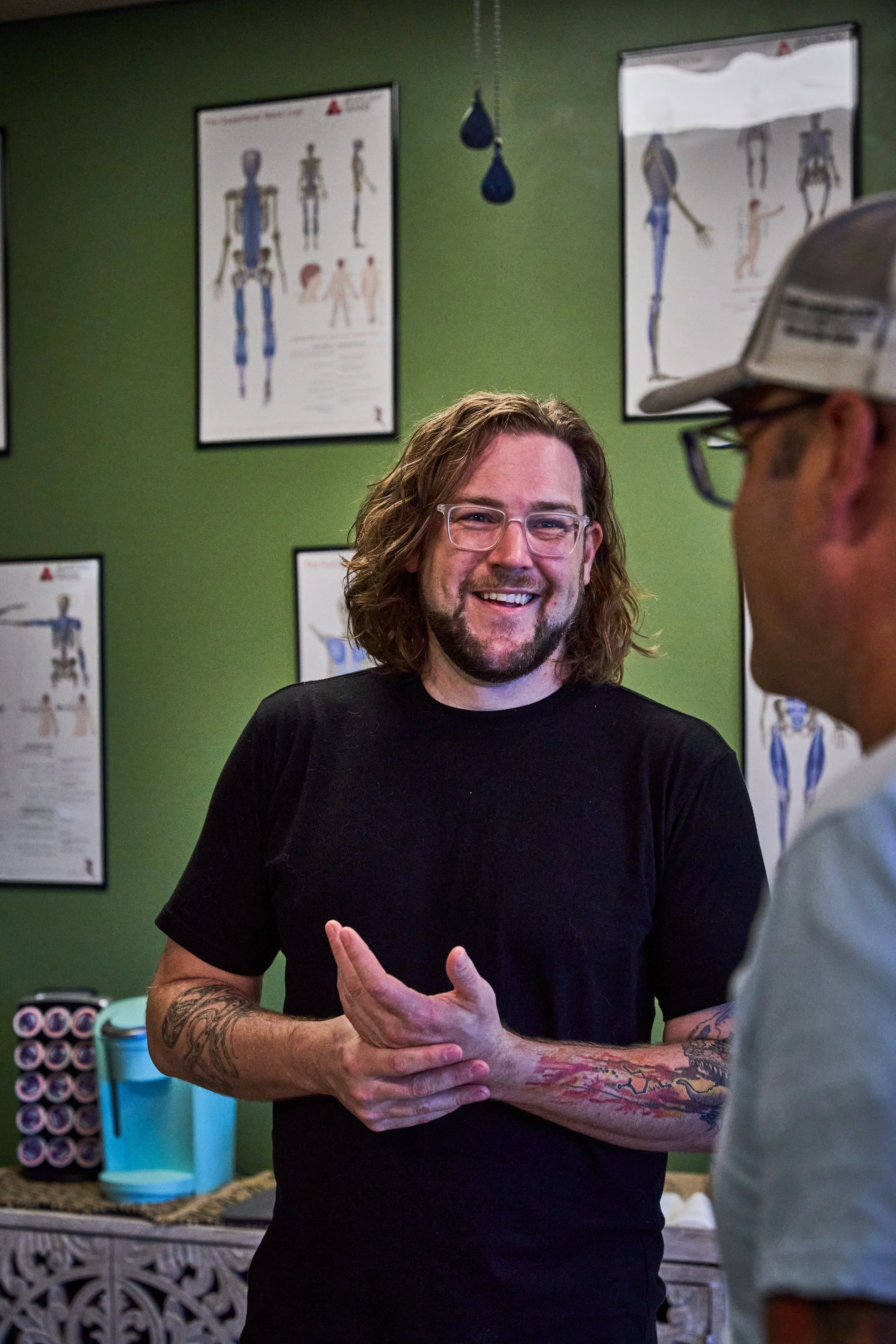 A man with wavy brown hair, glasses, and tattoos on his arms smiling and talking to another man in a cap inside a room with medical posters on a green wall.