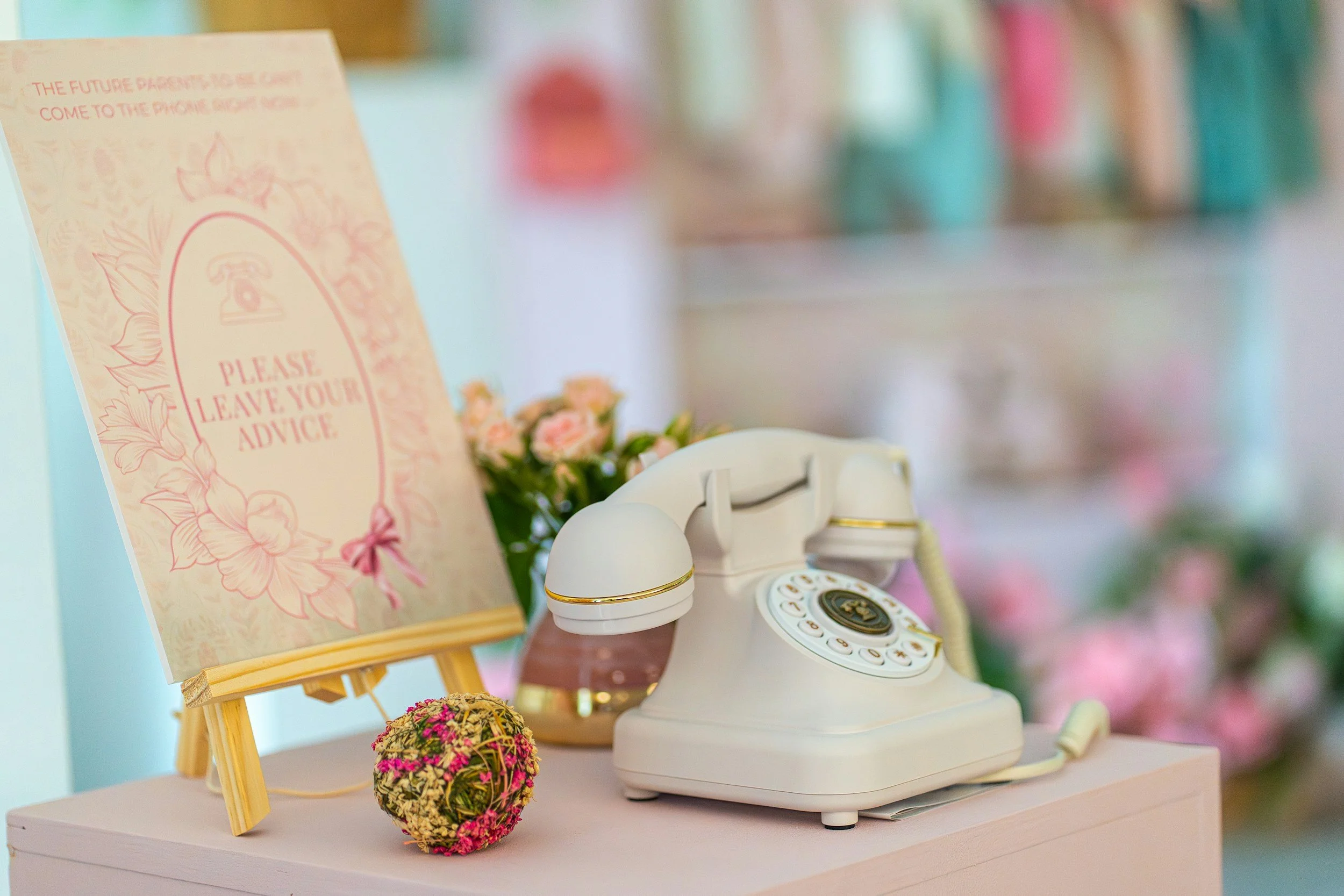 A white vintage rotary phone placed on a pink surface next to a small decorative ball of pink and green flowers, with a pink and white floral sign that reads "Please leave your advice" and a bouquet of pink flowers in the background.