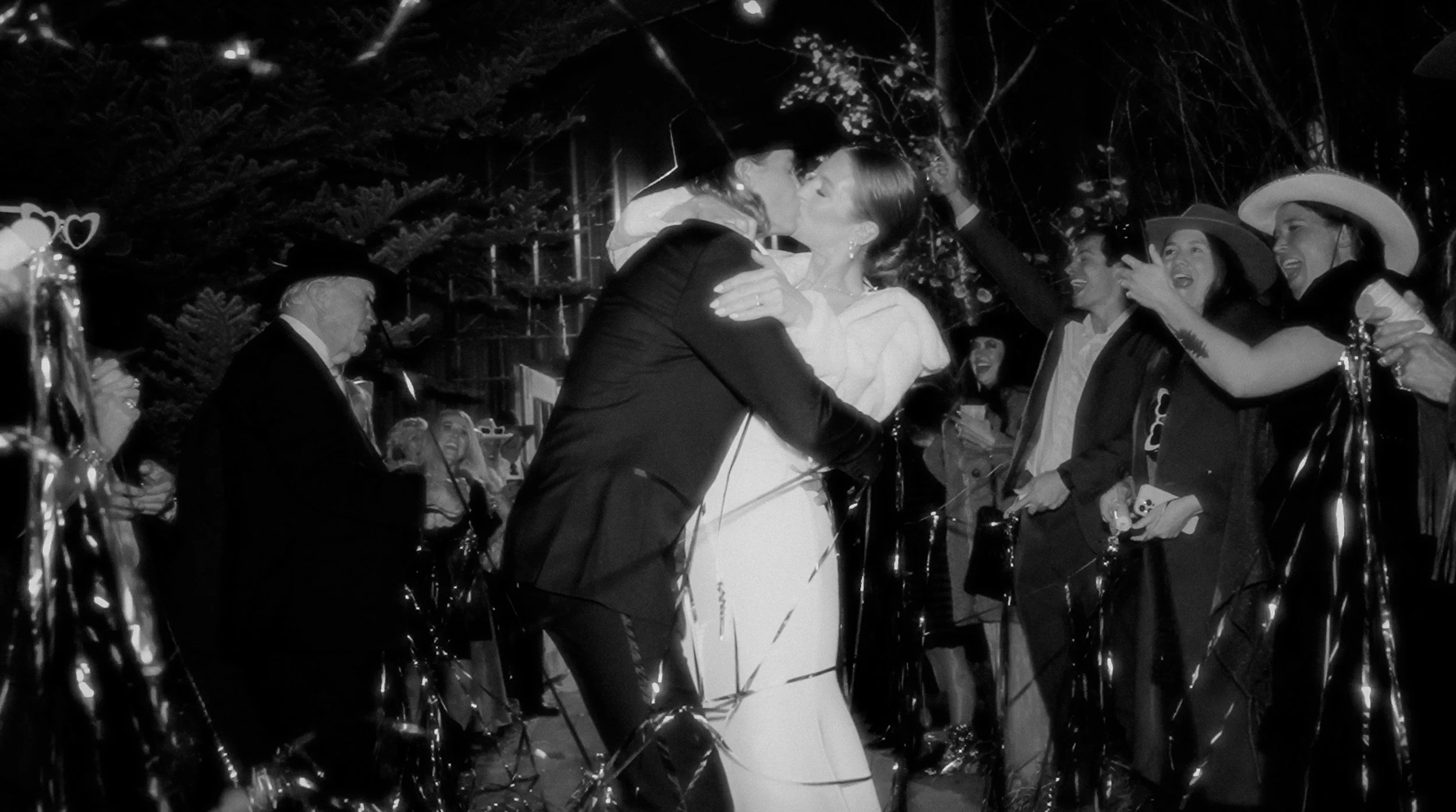 A couple in wedding attire kissing amid party celebration with friends and family, festive decorations, and string lights.