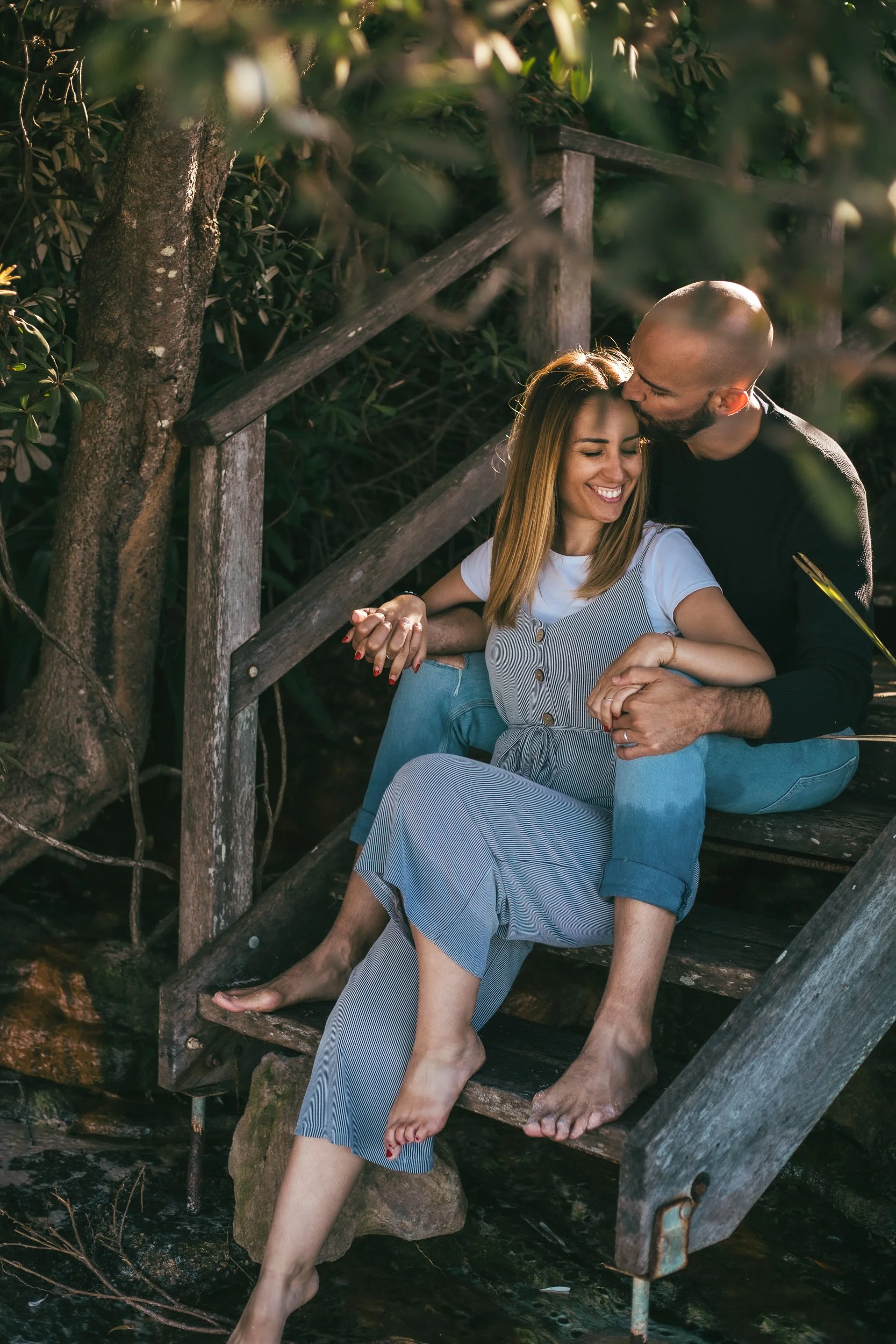 A couple sharing a kiss while sitting on a wooden staircase in a serene forest setting.
