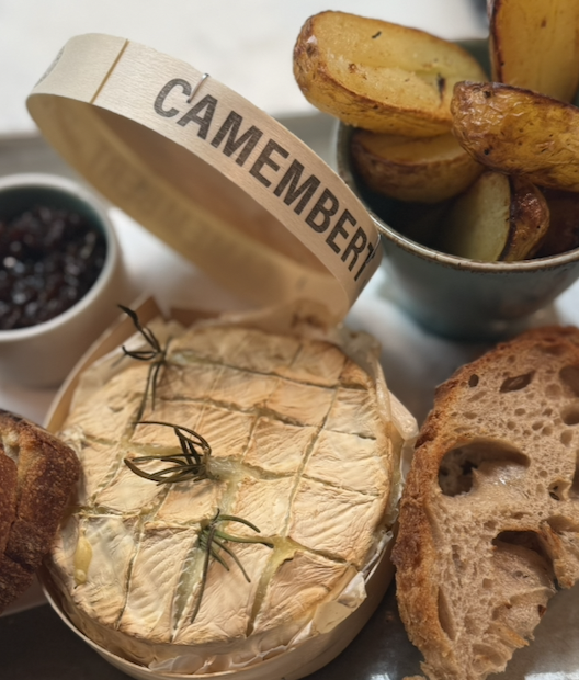 A wedge of camembert cheese garnished with herbs, a bowl of roasted potato wedges, slices of toasted bread, and a small cup of dark berry jam.