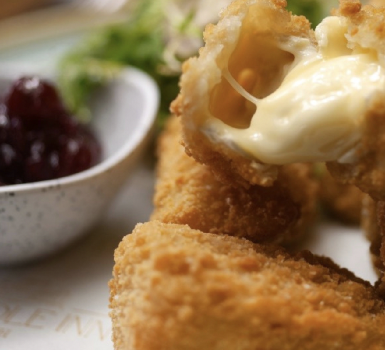 Close-up of a breaded fried chicken tender stuffed with melted cheese, with a side of berry jam and greens in the background.