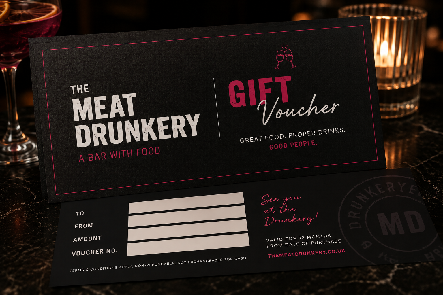 A gift voucher for The Meat Drunkery, a bar with food. The voucher is black with white and pink text, and background cups and glasses are visible.