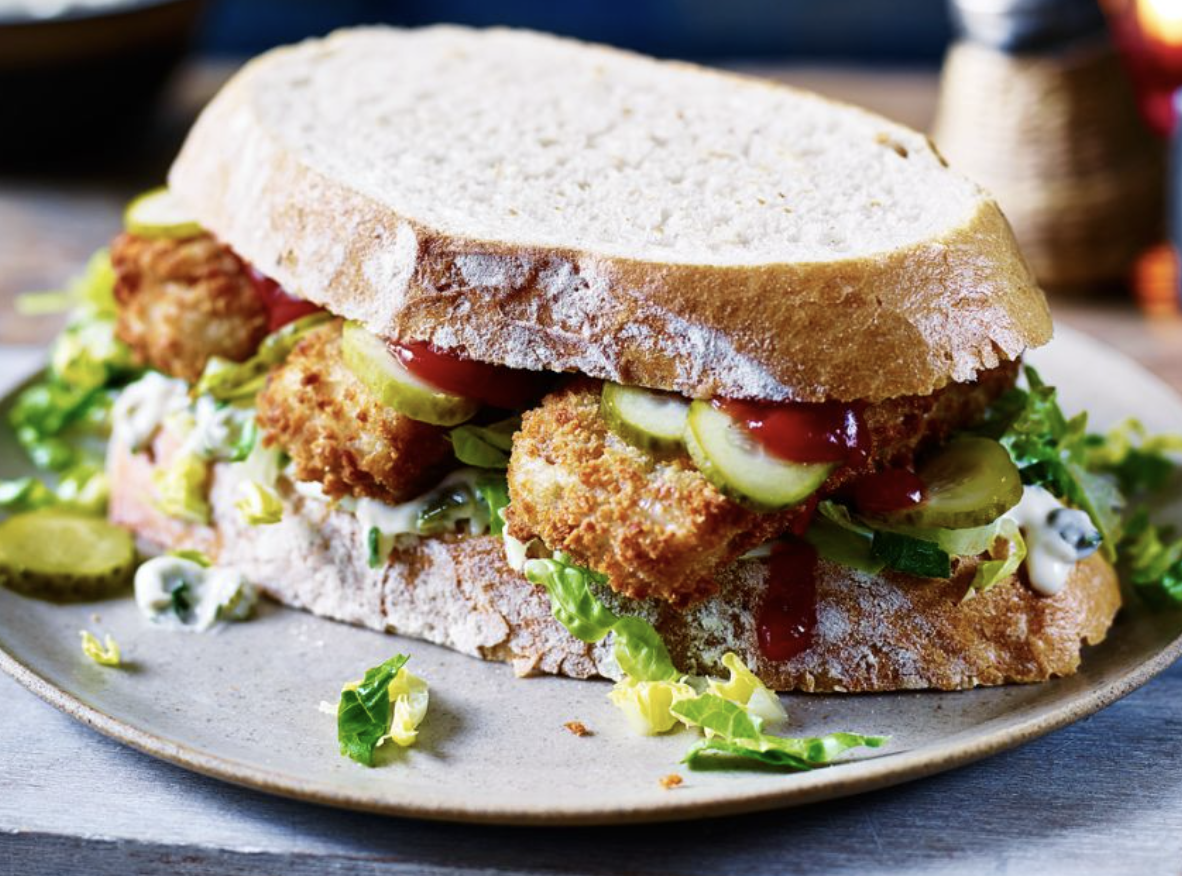 Fried fish sandwich with pickles, lettuce, tomato, mayonnaise, and ketchup on a white sandwich bun, served on a plate.