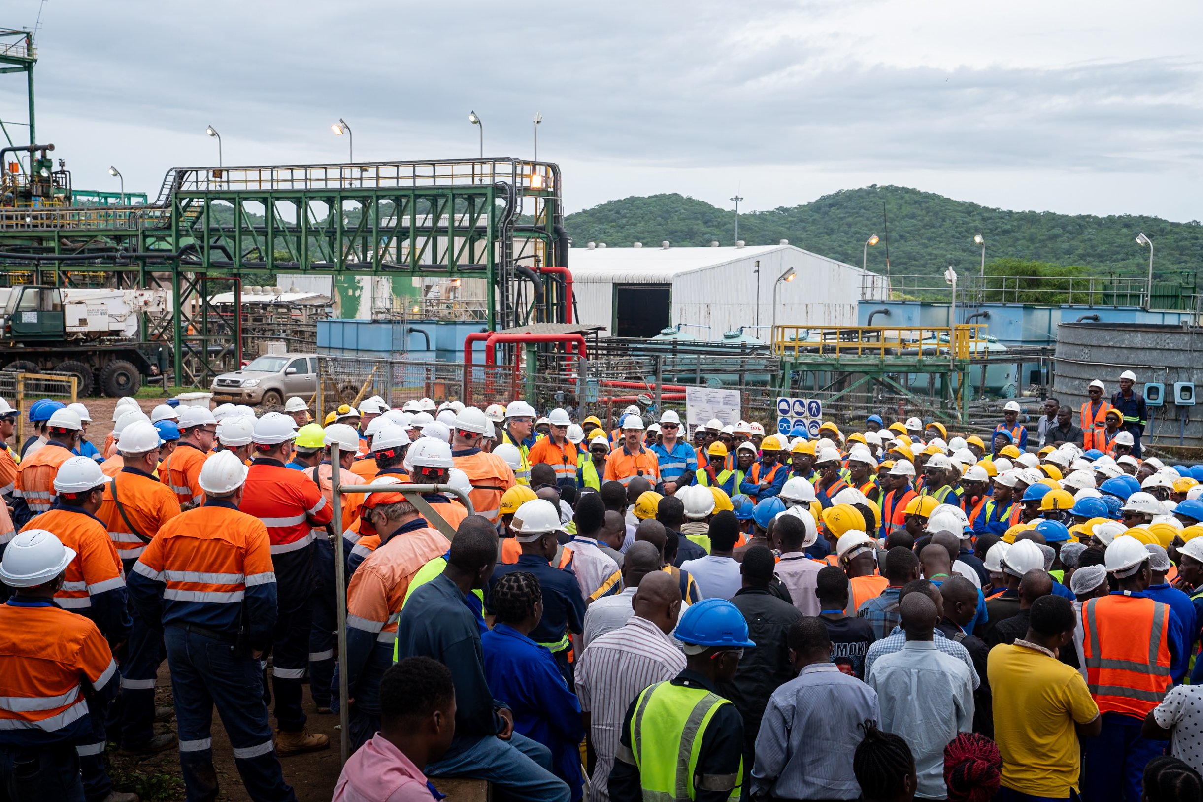 Large group of construction workers in safety gear gathered at an industrial site for a meeting with equipment and buildings in the background.