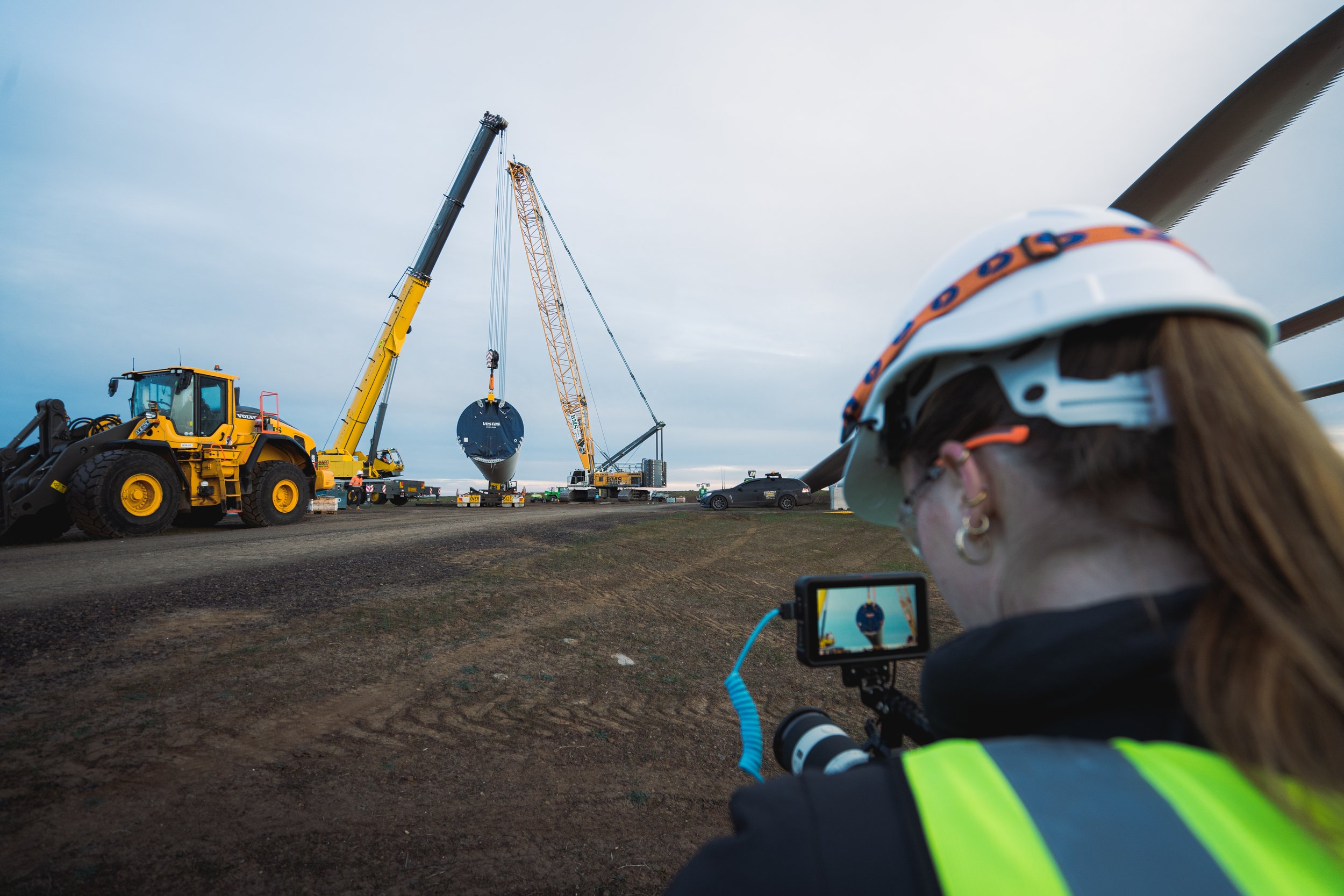 Construction site with cranes lifting a large cylindrical object, person in safety gear capturing image with camera.