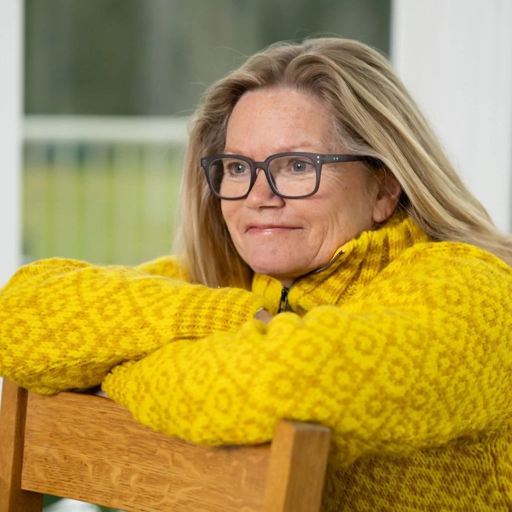 A woman with blonde hair and glasses, wearing a yellow patterned sweater, resting her arms on a wooden chair, smiling softly.