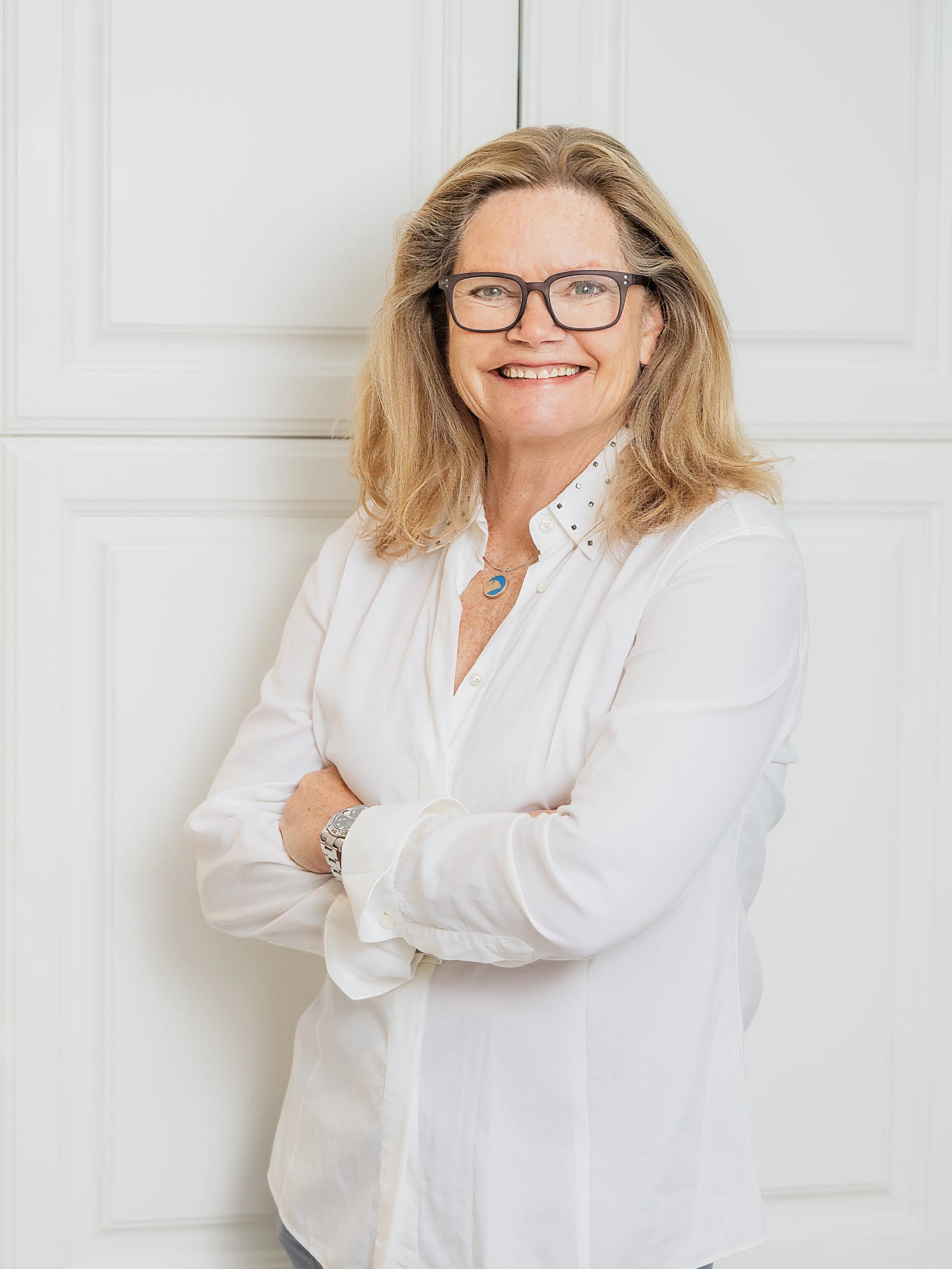 A smiling woman with glasses and shoulder-length blonde hair, wearing a white blouse, standing with arms crossed in front of a white paneled wall.