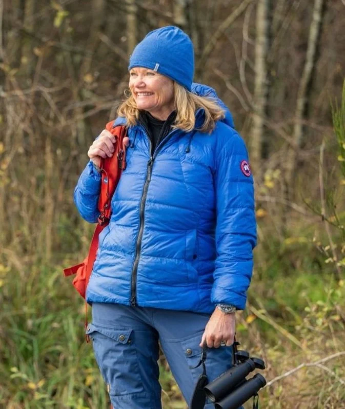 Woman wearing a blue jacket and matching beanie hiking outdoors with a red backpack and black binoculars, smiling among trees and bushes.