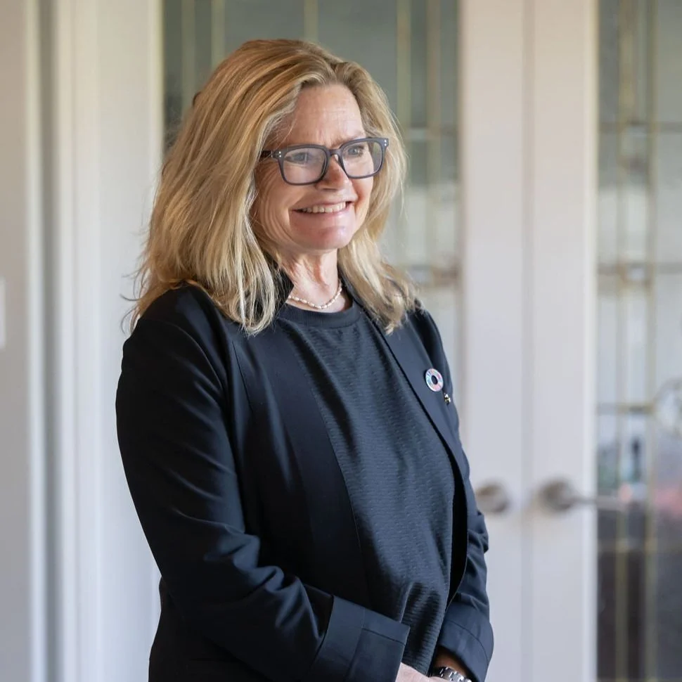 A woman with blonde hair, glasses, and a black blazer, smiling while standing indoors in front of glass doors.