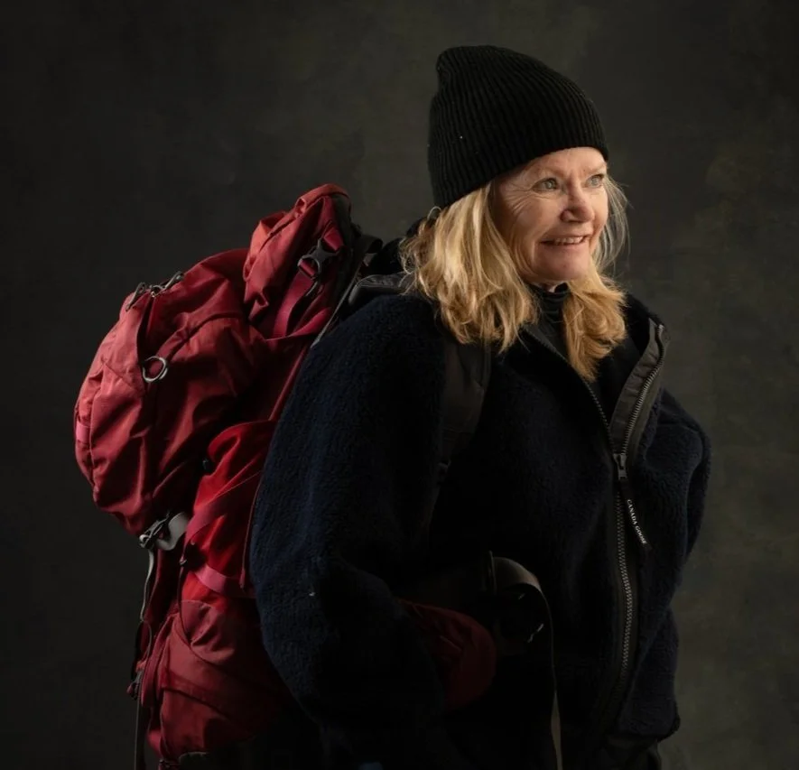 Woman with blonde hair wearing a black beanie, black fleece jacket, and backpack, smiling at the camera, against a dark background.