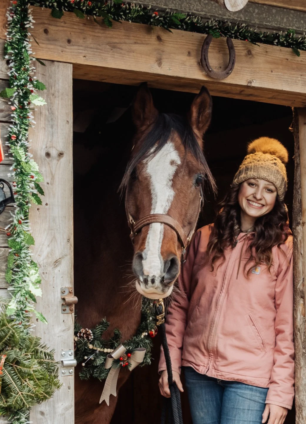 A young woman standing next to a brown horse decorated with Christmas garland inside a stable. The woman is wearing a tan knit hat with a pom-pom, a light pink jacket, and smiling at the camera.