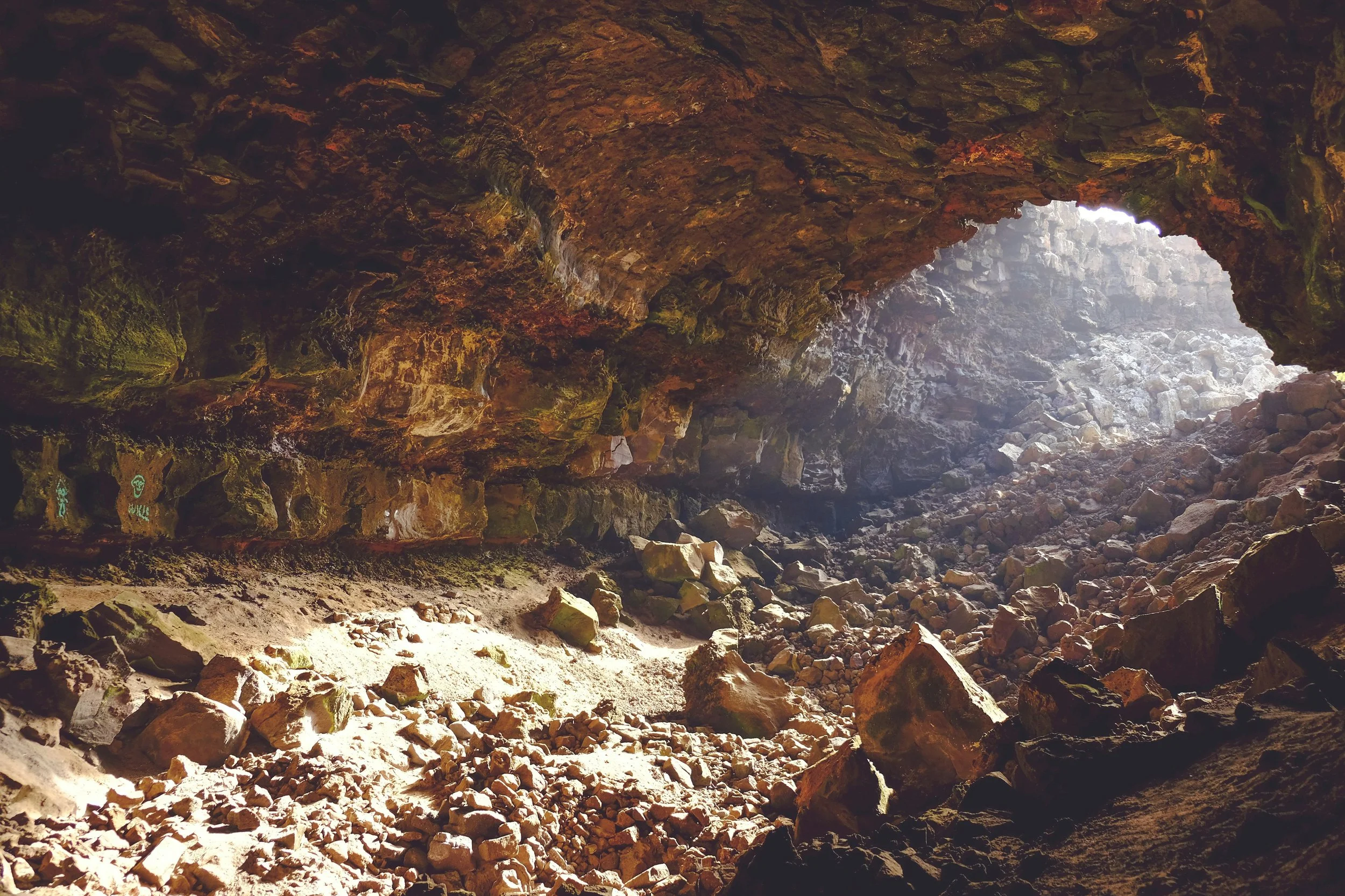 Inside a rocky cave with a large opening letting in sunlight, revealing rocks and dirt on the cave floor with moss-covered rocks on the walls.