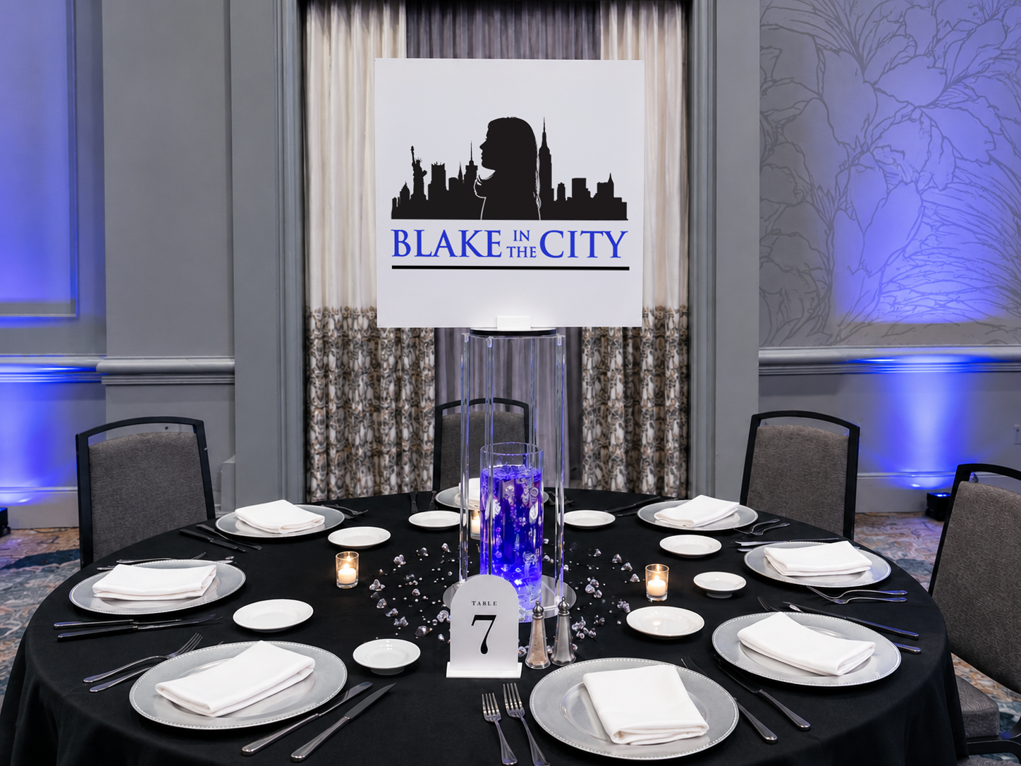 Round banquet table with black tablecloth, white plates, cloth napkins, and silverware, set for eight. The centerpiece features a tall, clear cylinder filled with decorative blue water and illuminated from below, with a large sign above displaying the words 'Blake in the City' and a silhouette of a woman's profile against a city skyline. There are small candles and scattered crystals on the table, and a table number card indicating 'Table 7.' The background includes patterned curtains and wall decor with blue lighting accents.