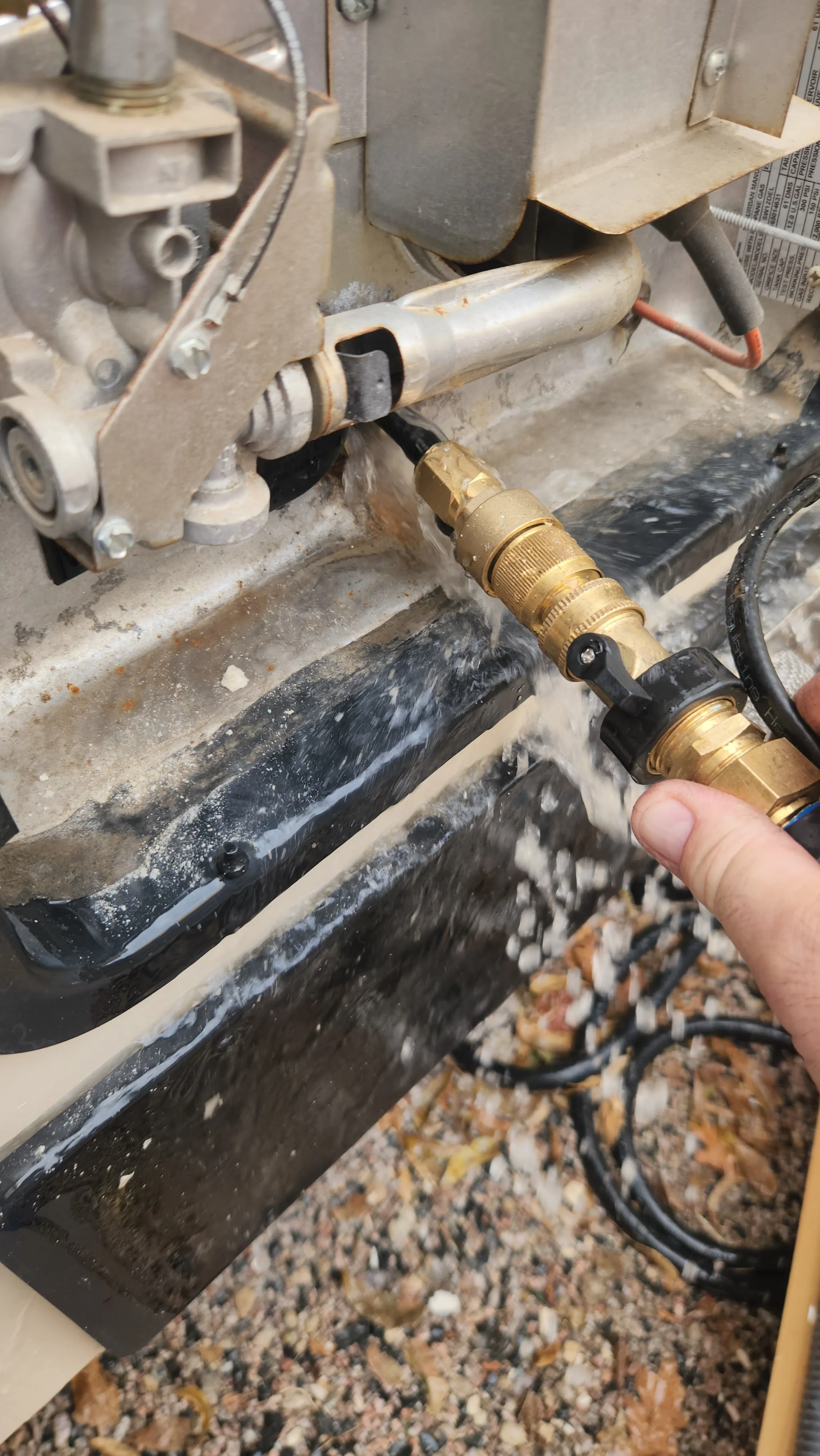 A person is using a pressure washer to clean a machine or piece of equipment outdoors. The pressure washer has a brass nozzle, and water is being sprayed onto the equipment, which appears to have some dirt and grime.