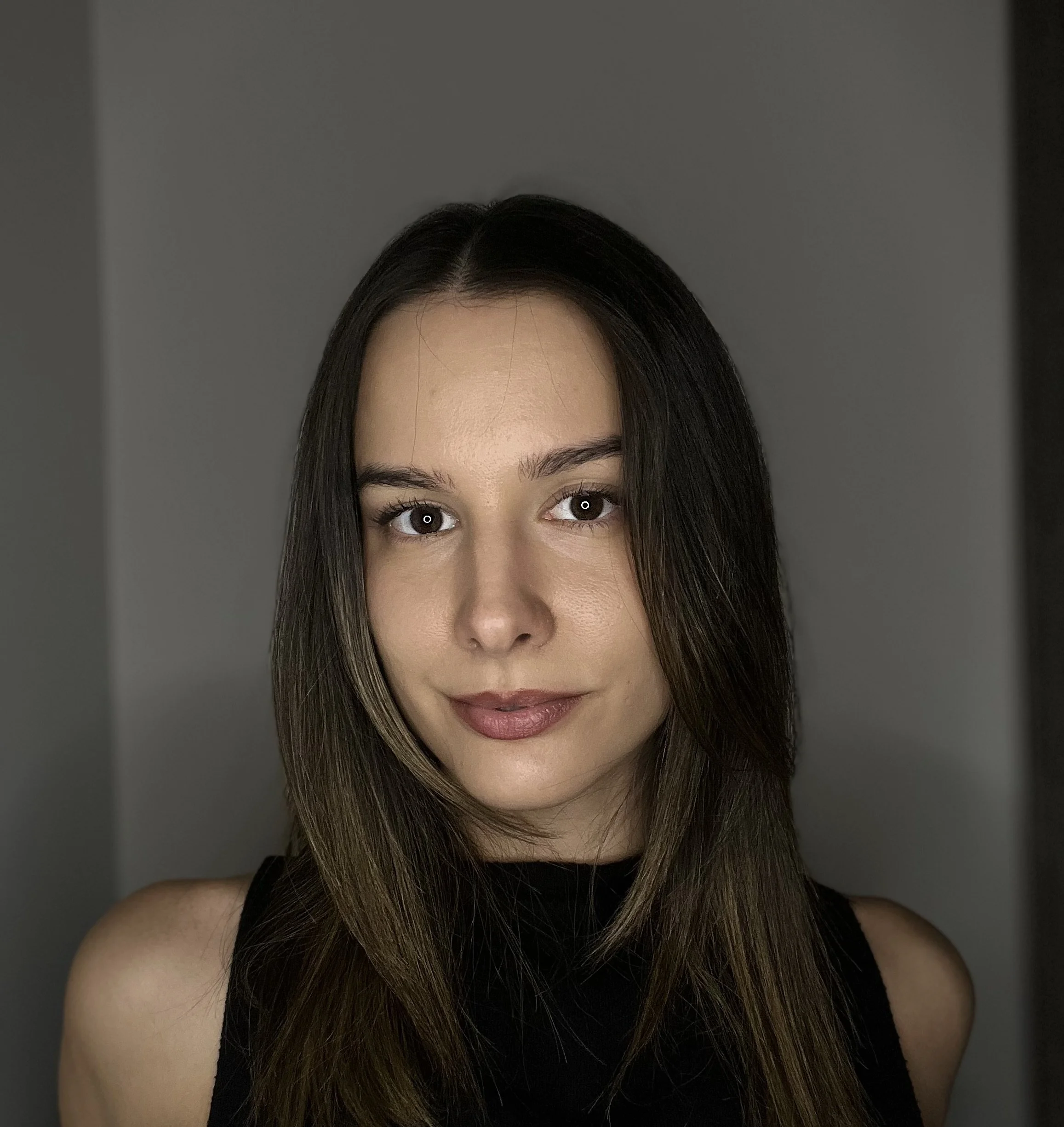 Close-up of a woman's face with brown eyes, dark eyebrows, and long dark hair, wearing a black top, against a gray background.