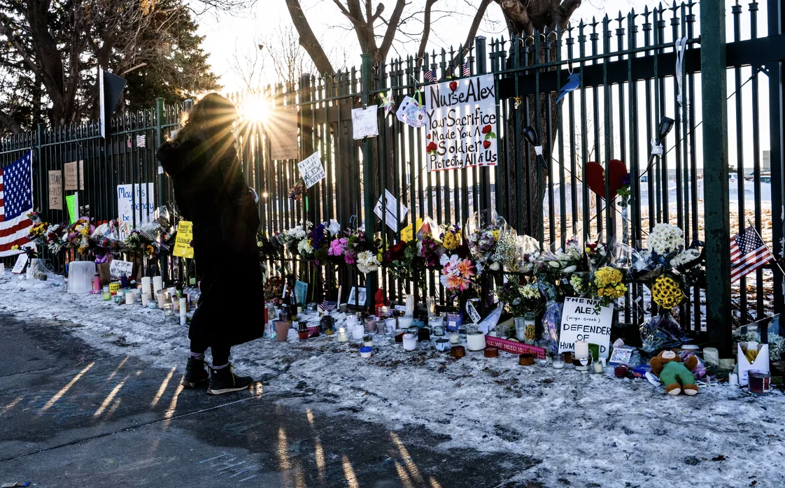 woman stands at vigil on sidewalk, looking at flowers and memorials