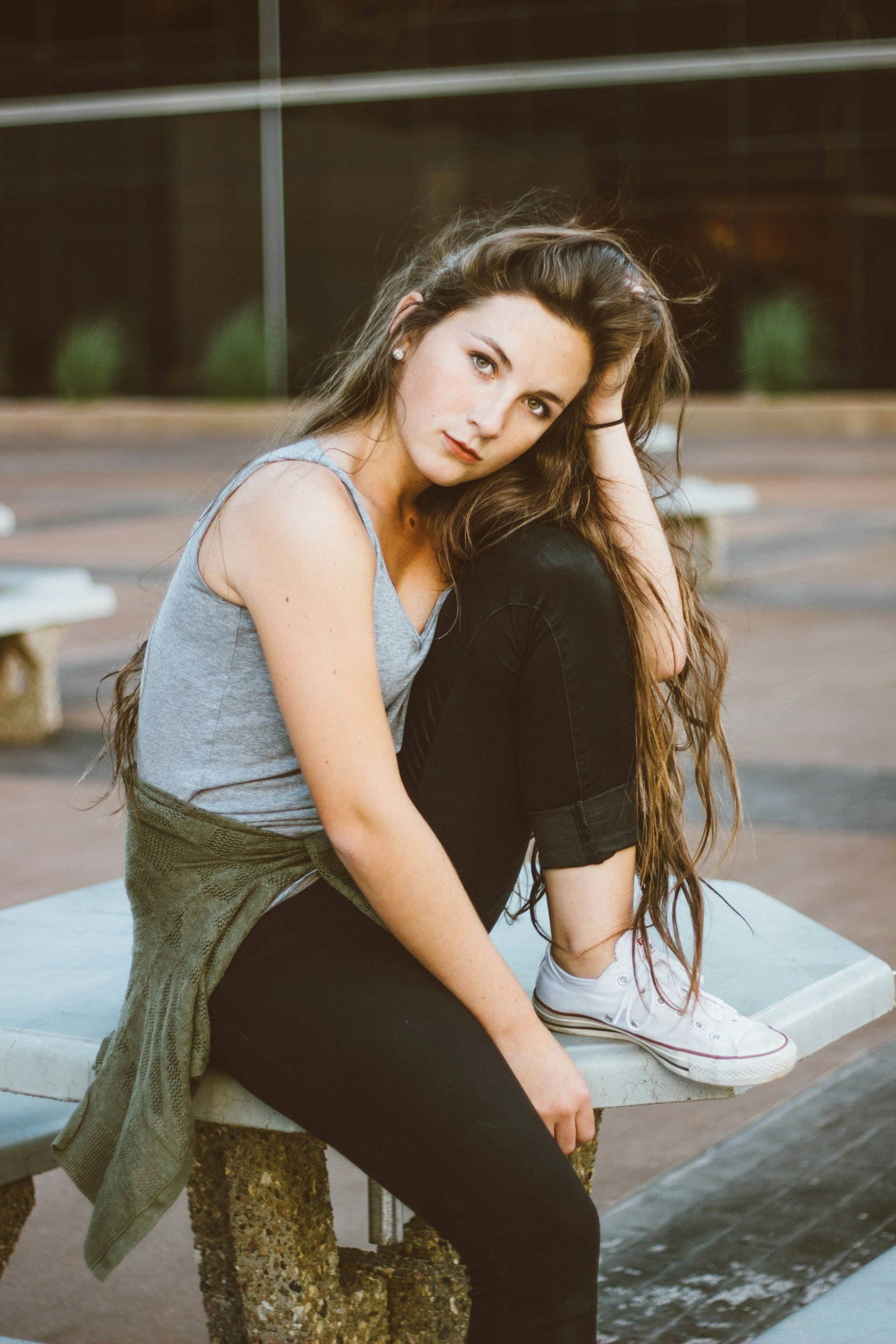 A young woman with long brown hair and light skin sits on a concrete ledge in an outdoor setting, with her elbow on her knee and her head resting on her hand. She is wearing a gray tank top, black pants, and white sneakers, and looks at the camera with a neutral expression.
