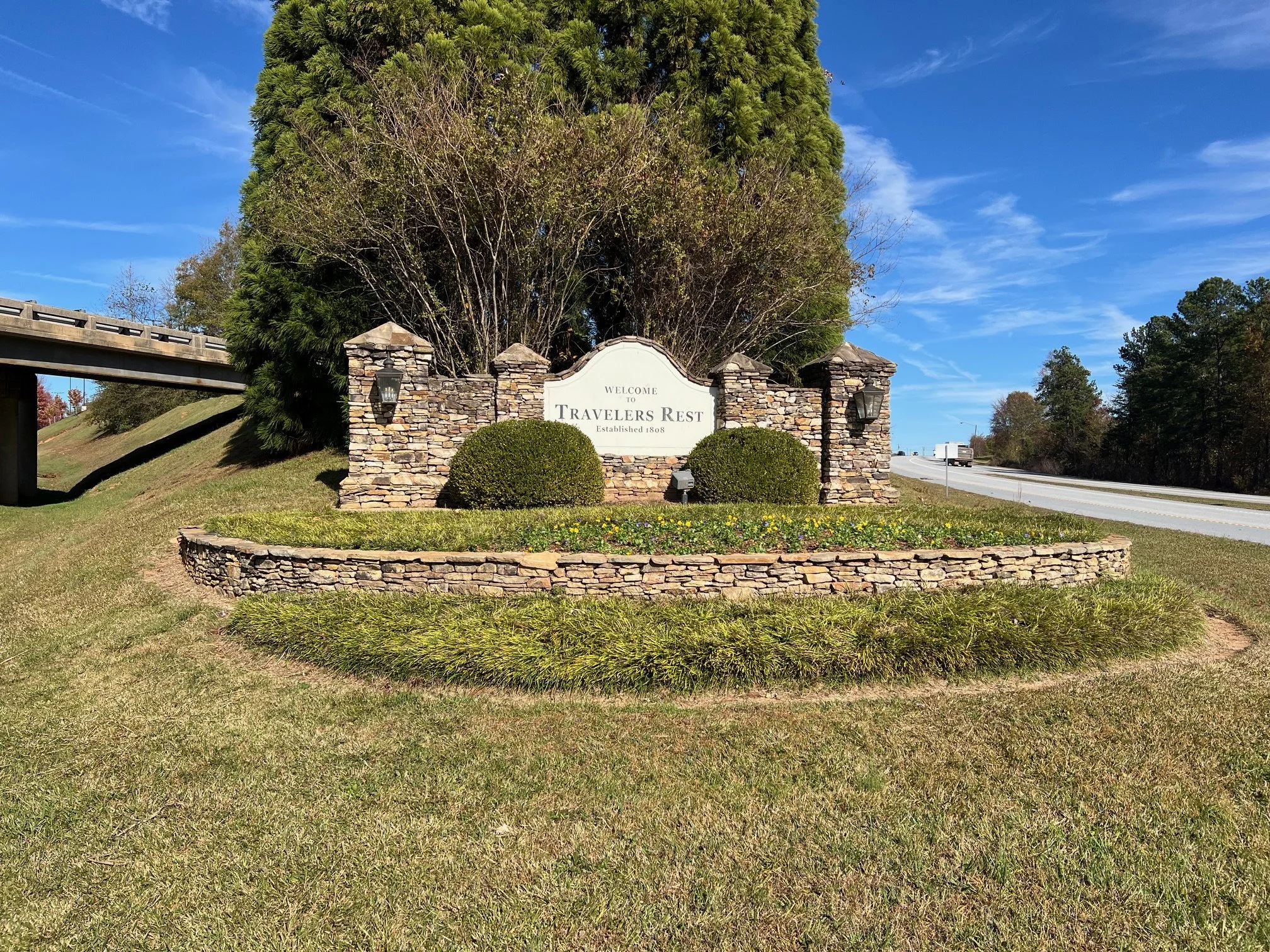 Entrance sign for Travelers Rest, surrounded by a stone wall with greenery and trees in the background under a blue sky.