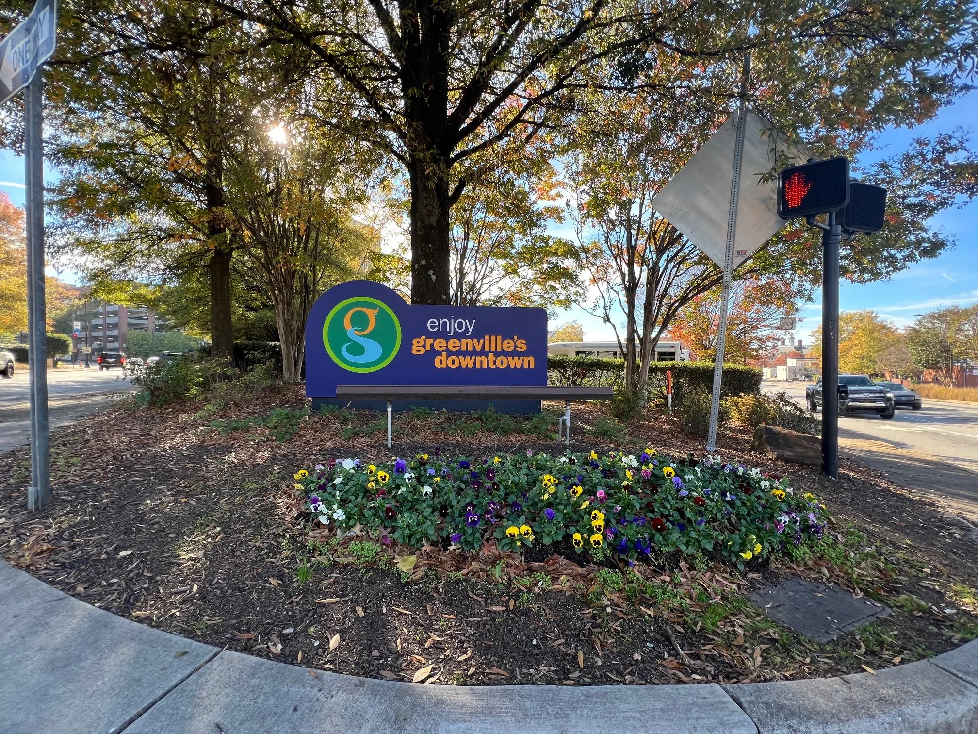 A sign welcoming visitors to Greenville's downtown, surrounded by a bed of colorful pansies, under trees with autumn foliage, near a busy street with cars and traffic signals.