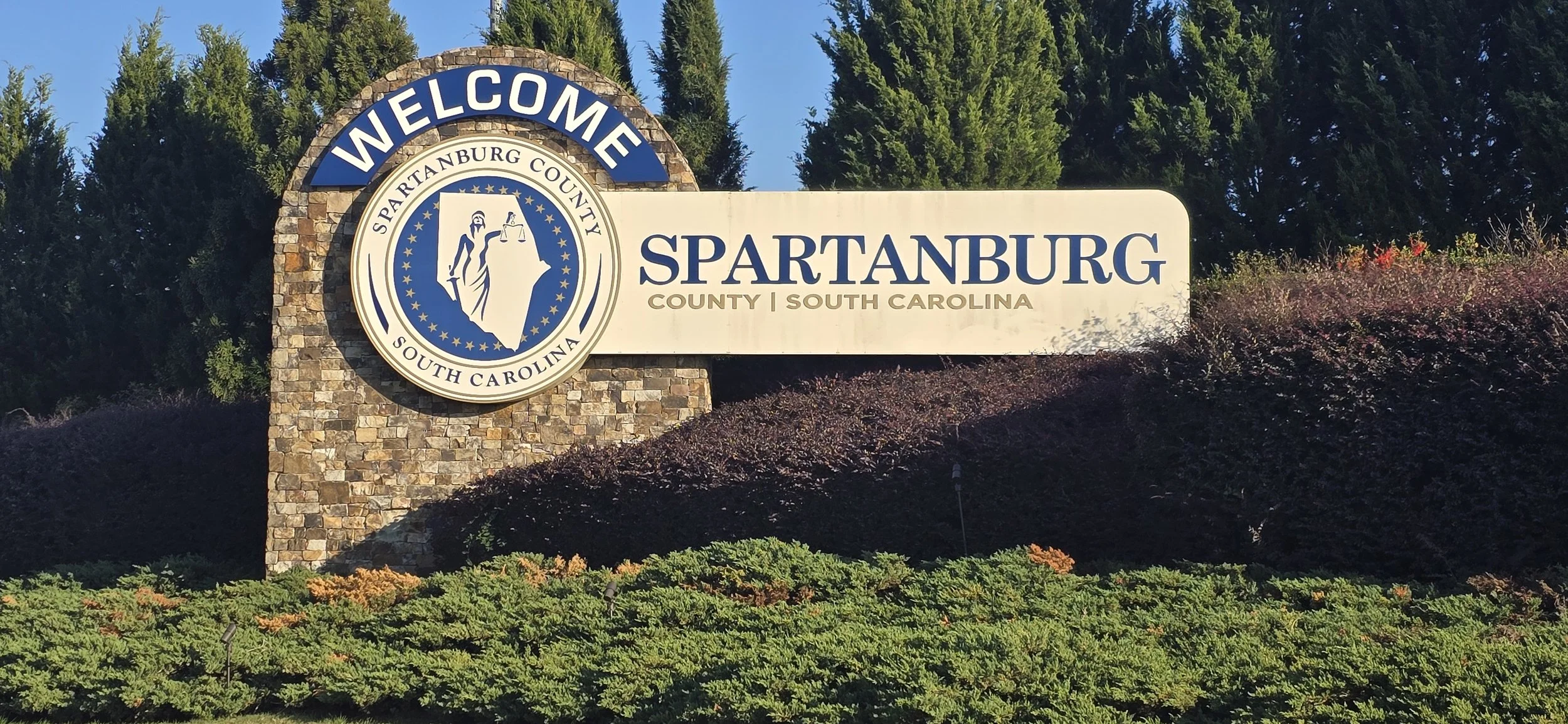 Welcome sign for Spartanburg County, South Carolina, with a circular emblem of Lady Justice holding scales, surrounded by trees and bushes.