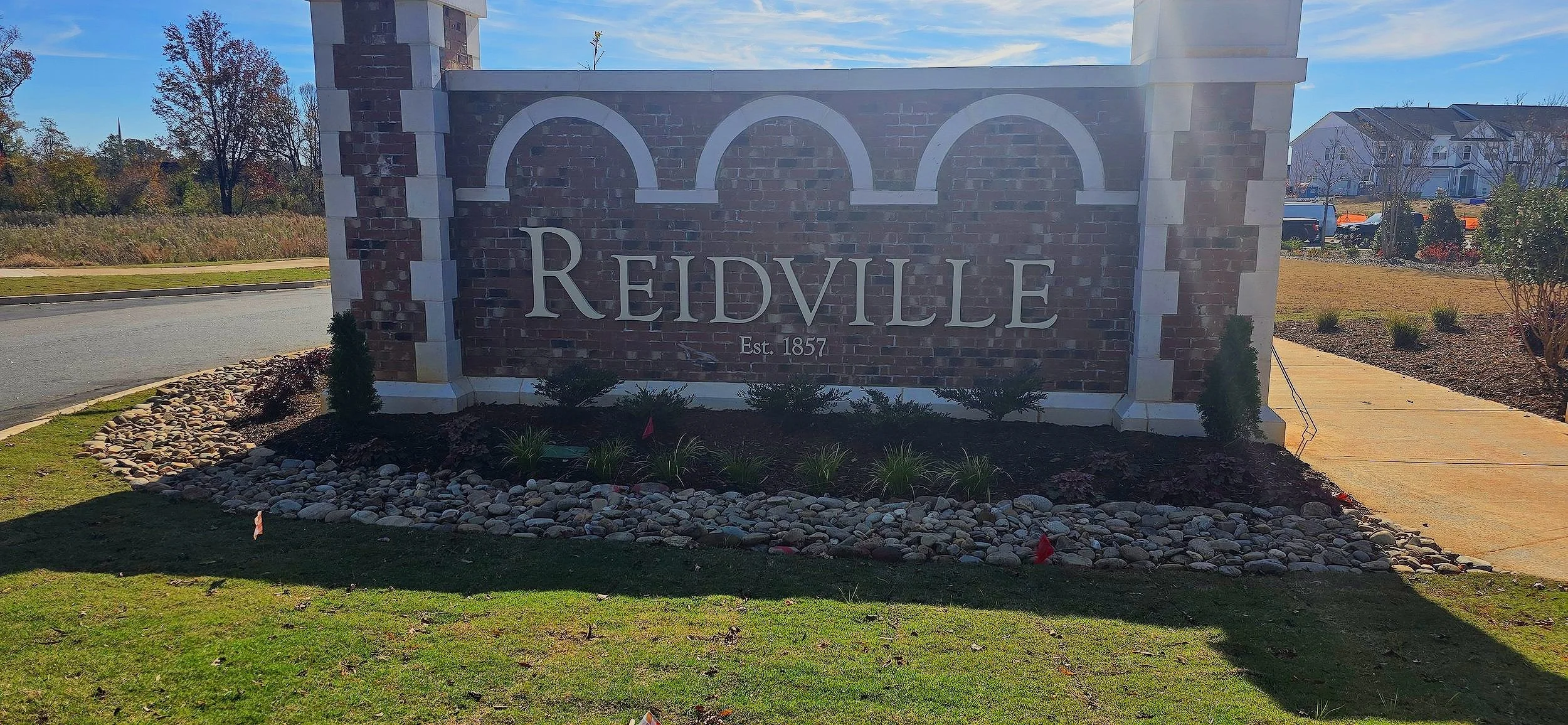 A brick and stone sign that reads 'REIDVILLE Est. 1857' with a landscaped area in front and a sidewalk to the right, under a clear blue sky.