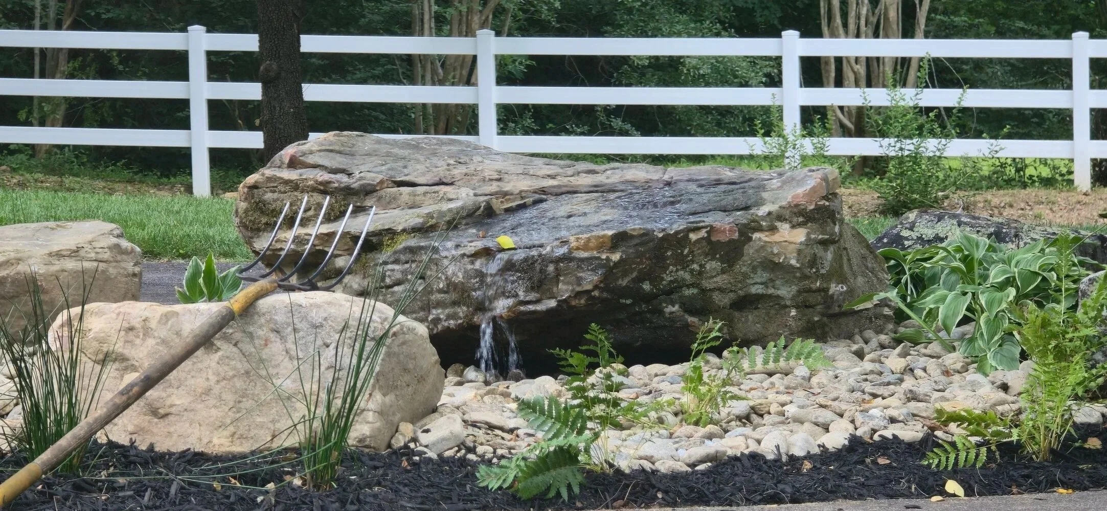 A garden landscape with a waterfall feature, large rocks, green plants, and a white fence in the background.