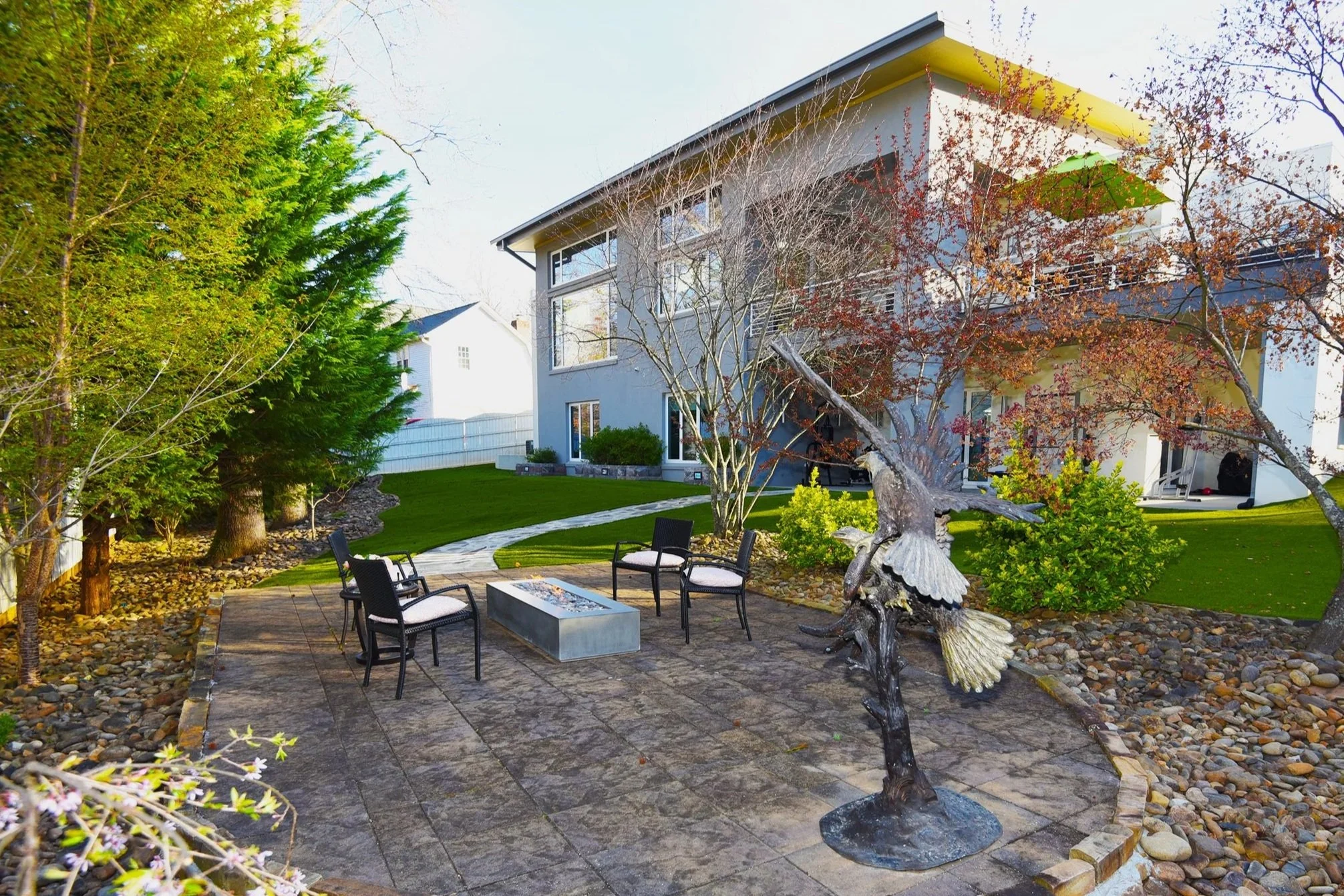 The event space extends toward the modern home, with the fire pit, sculpture, and surrounding plantings creating a harmonious connection between landscape and architecture.