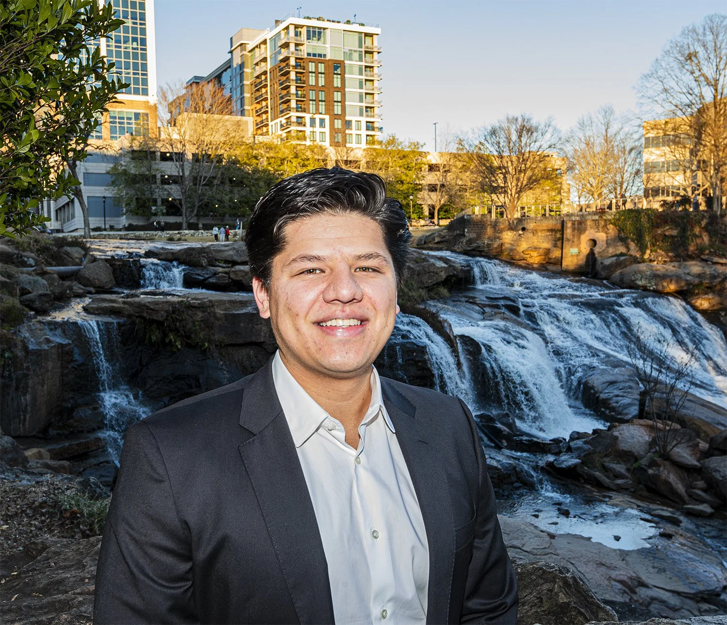 A man in a suit smiling outdoors near a waterfall with city buildings in the background.