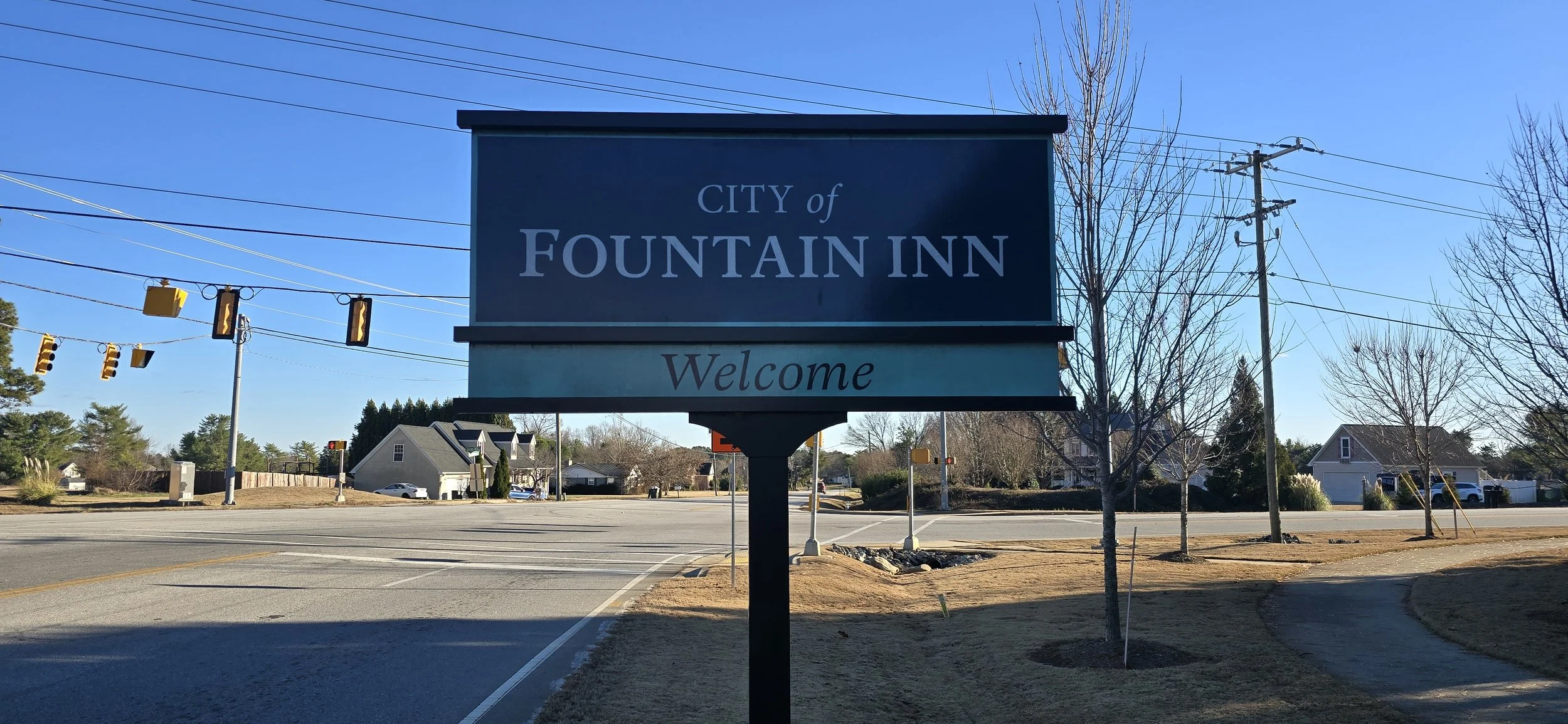Sign reading 'City of Fountain Inn Welcome' in a suburban area with trees, houses, and traffic lights in the background on a clear day.