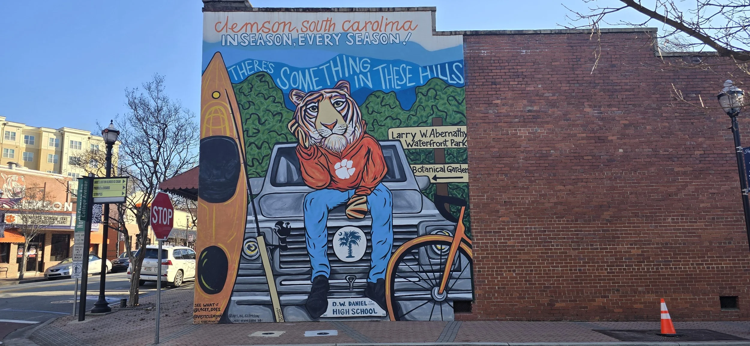 Colorful mural in Clemson, South Carolina, depicting a tiger mascot sitting on a car, holding a paddle, with a kayak and a bicycle nearby. The mural has text saying 'There's something in these hills' and references local parks and a high school.
