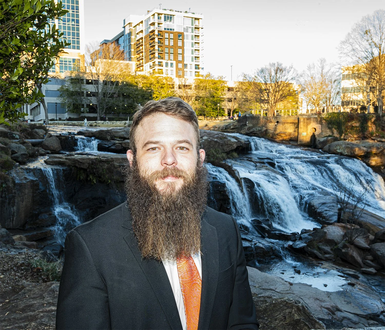 A man with a full beard wearing a black suit and an orange tie standing outdoors near a waterfall in an urban park, with modern buildings and trees in the background during the daytime.