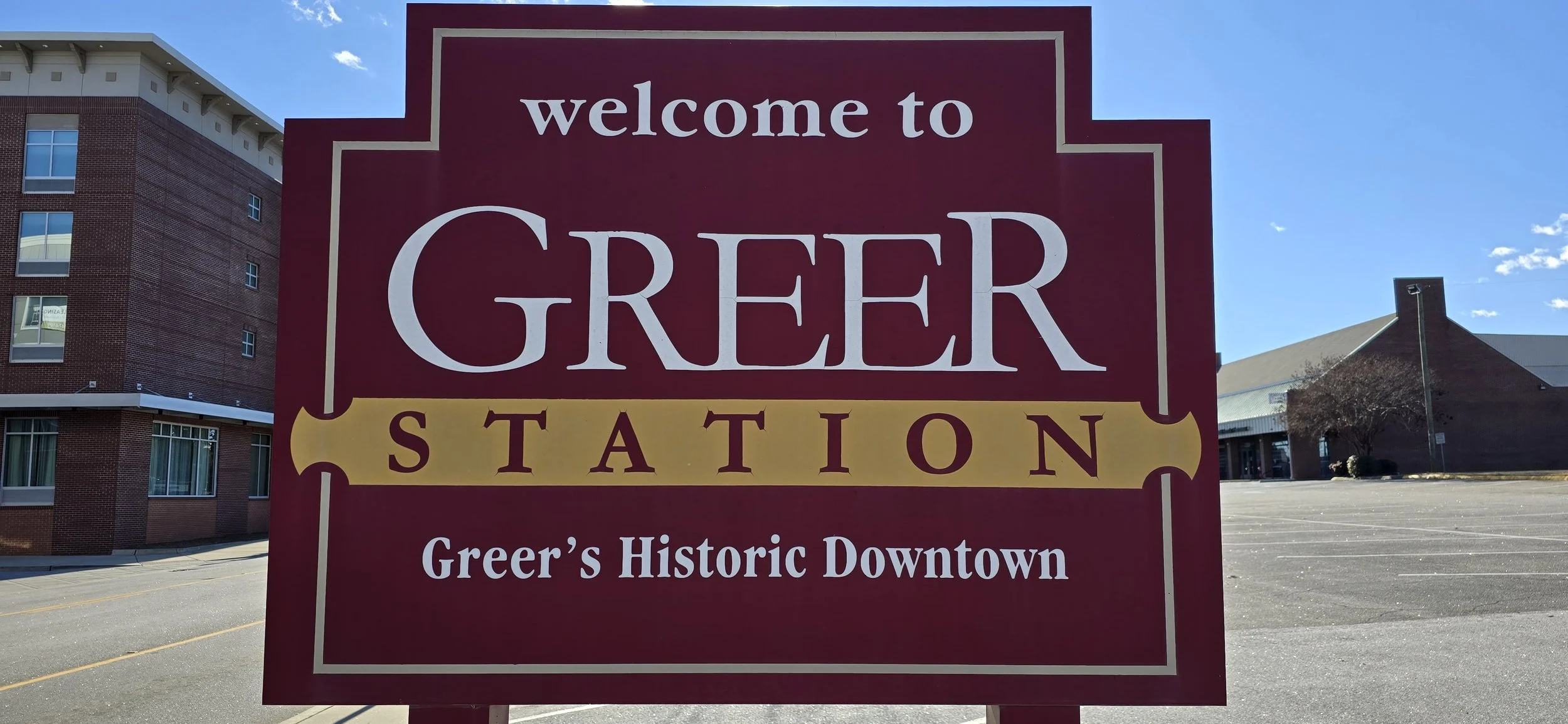 A large public sign saying "welcome to GREER STATION Greer's Historic Downtown" in front of a parking lot and buildings in Greer, South Carolina, on a sunny day.