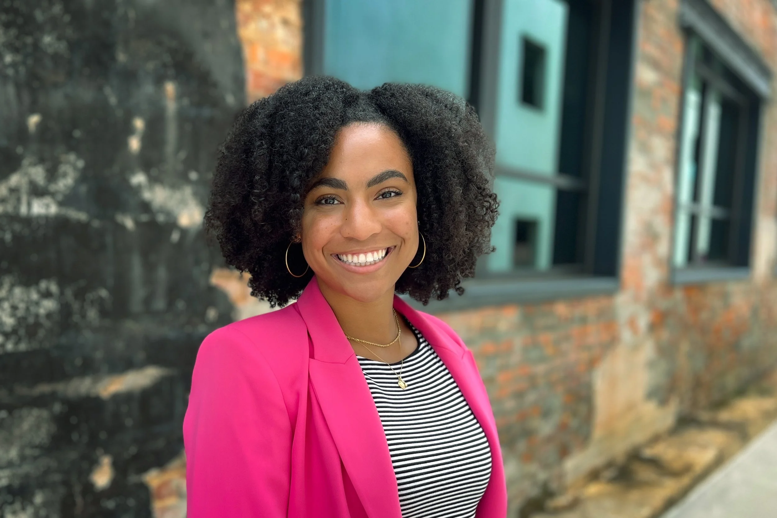 Portrait of a woman with curly black hair, wearing a bright pink blazer, black and white striped top, gold jewelry, smiling, standing outdoors in front of a brick and blue building.