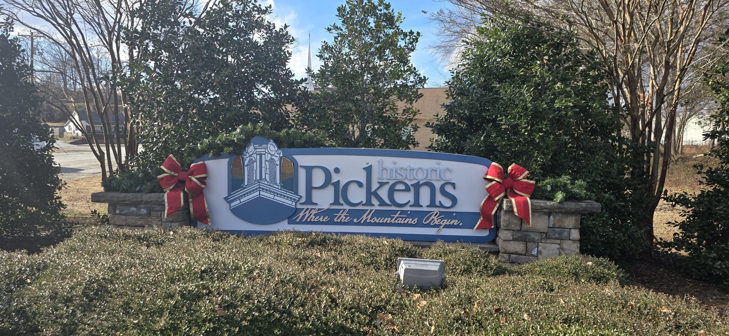 A sign for Pickens, South Carolina, reading "Historic Pickens - Where the Mountains Begin," decorated with holiday bows and greenery, surrounded by bushes and trees.
