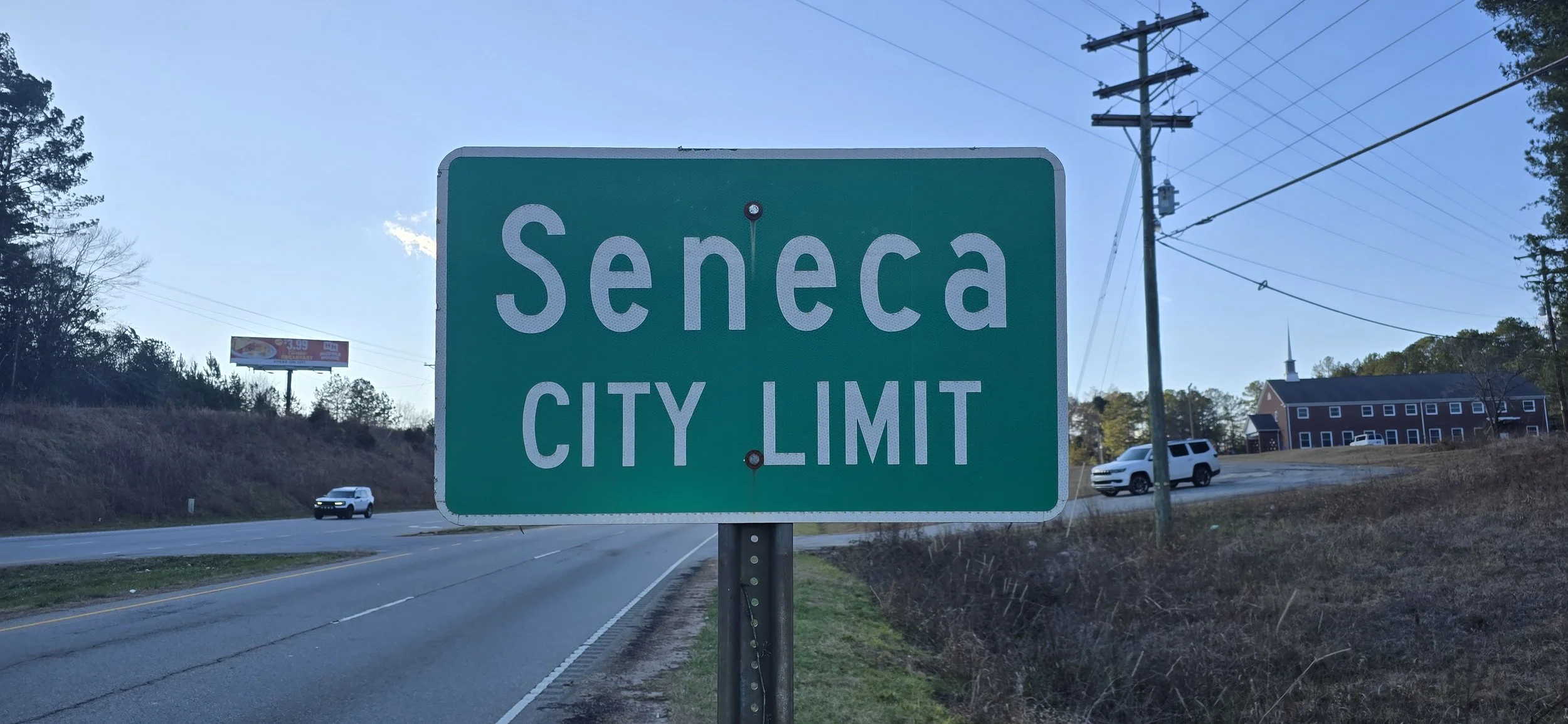 Green road sign that reads 'Seneca City Limit' along a highway with a white truck and a white SUV passing by, power lines, and a building with a steeple in the background.