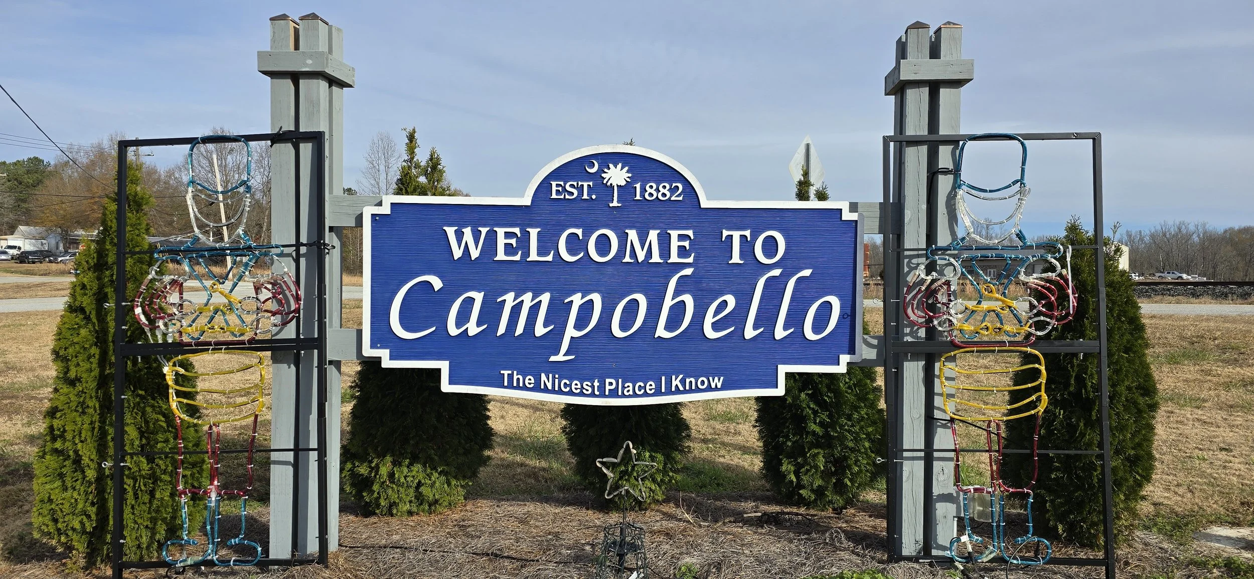 Blue welcome sign for Campobello with white text, flanked by decorative wire sculptures of colorful human figures, outdoors with trees and a cloudy sky.